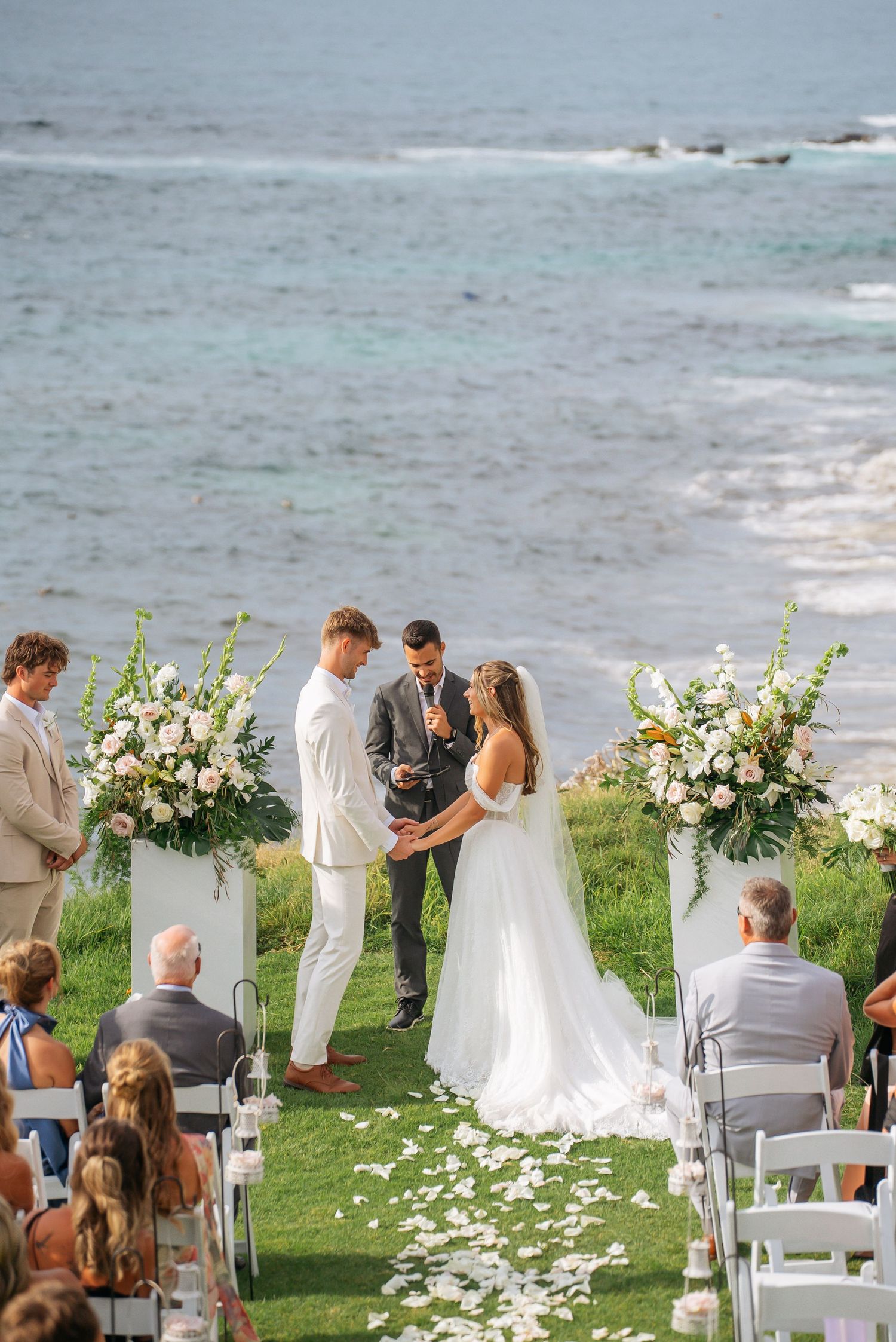 Outdoor oceanside wedding ceremony with couple exchanging vows flanked by white floral arrangements.