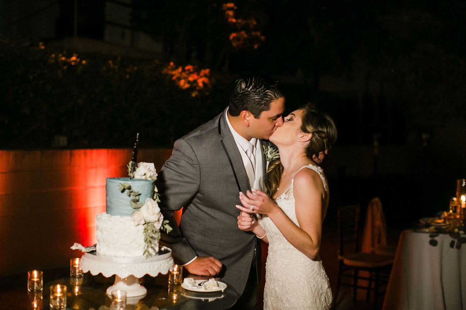 Bride and groom share a kiss while cutting their white wedding cake at an outdoor evening reception.