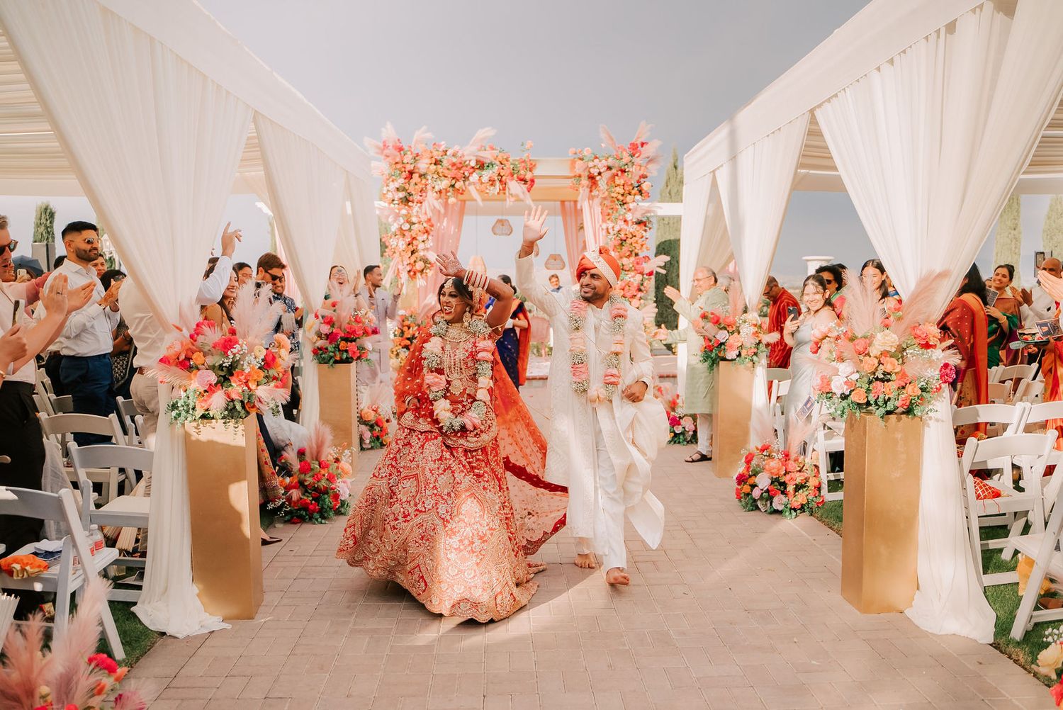 Indian bride and groom walk down aisle under floral mandap at elegant outdoor Hindu wedding ceremony.