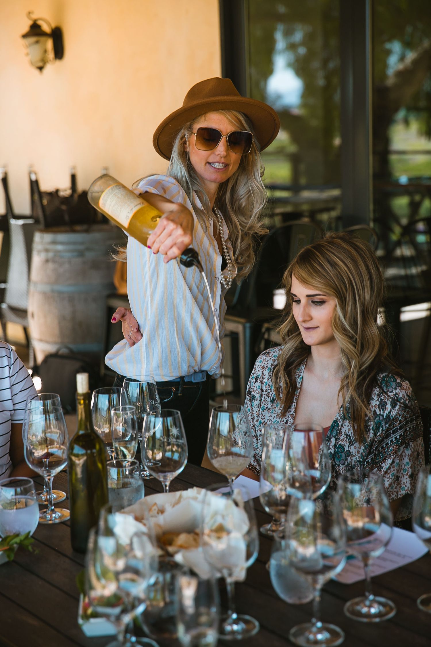 Woman in hat pouring white wine at outdoor winery table with guests and wine glasses during tasting.