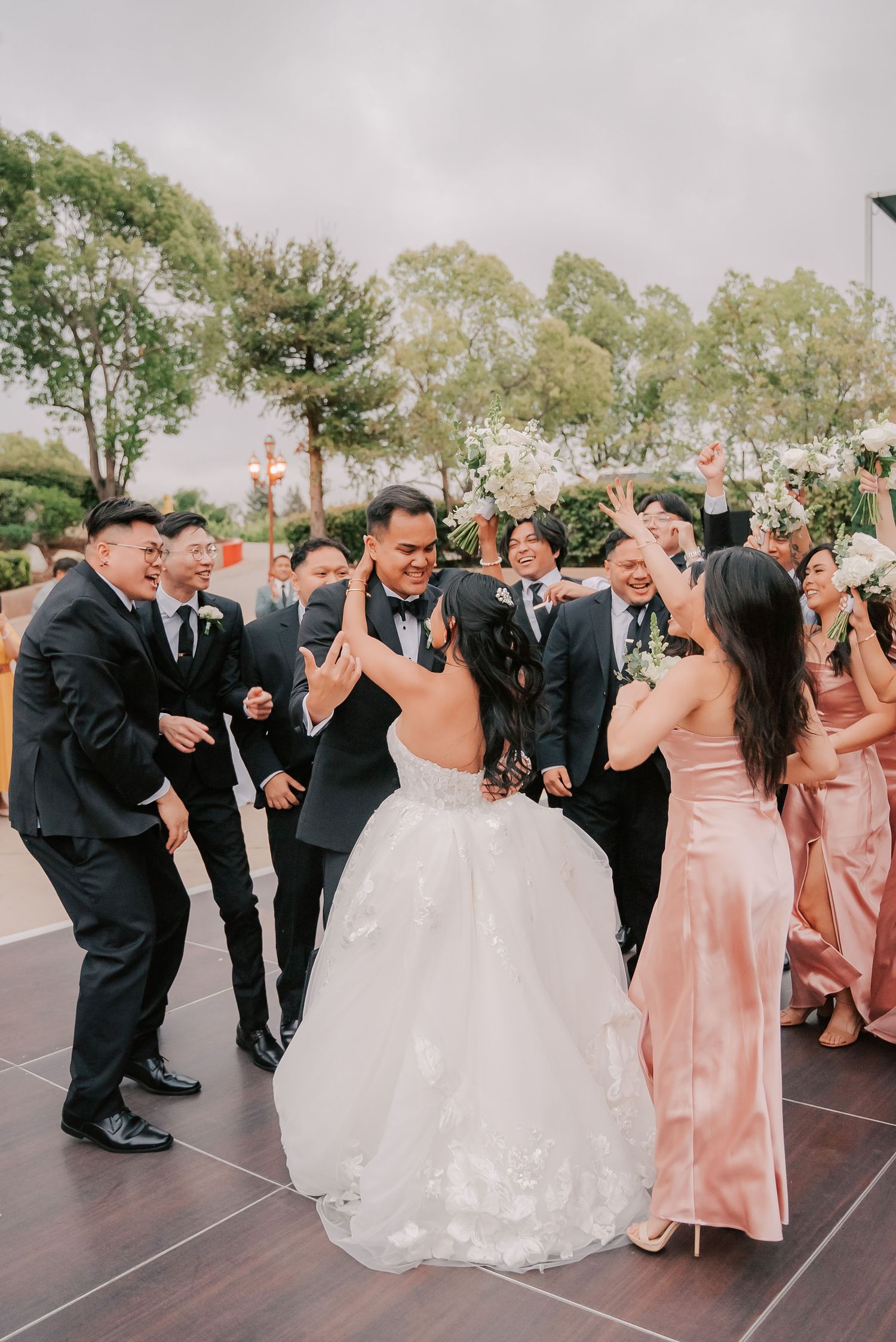Bride and groom celebrate with wedding party outdoors, guests cheering in black suits and pink bridesmaid dresses.