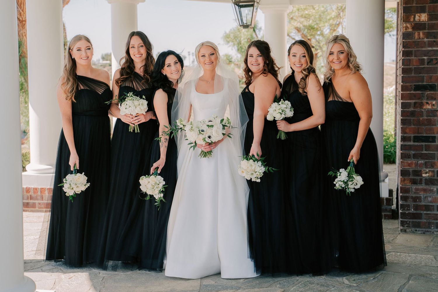 Bride in white gown poses with six bridesmaids in black dresses holding white floral bouquets at elegant venue.