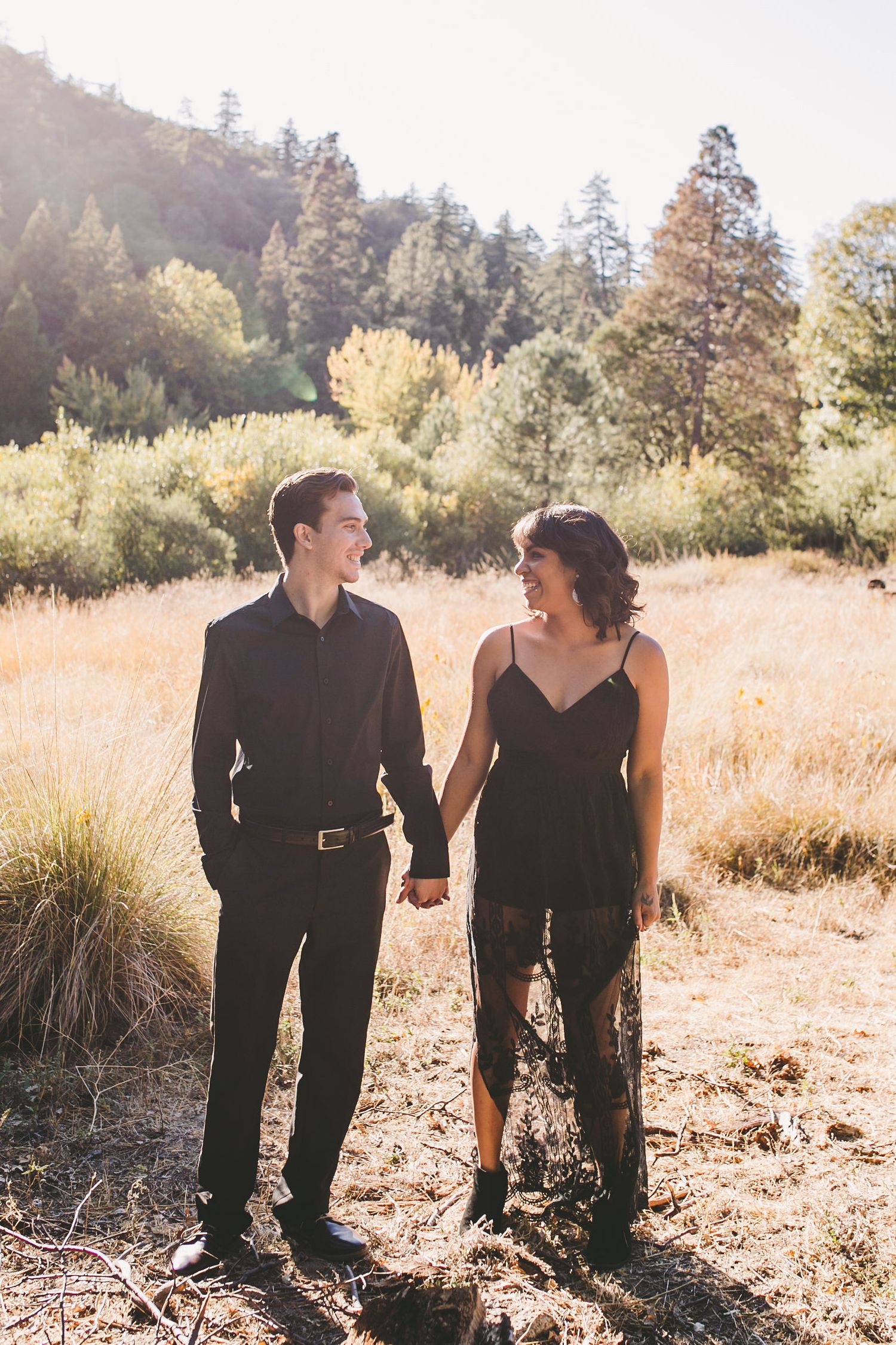 Couple in matching black outfits holding hands in a sunlit meadow with trees in the background.
