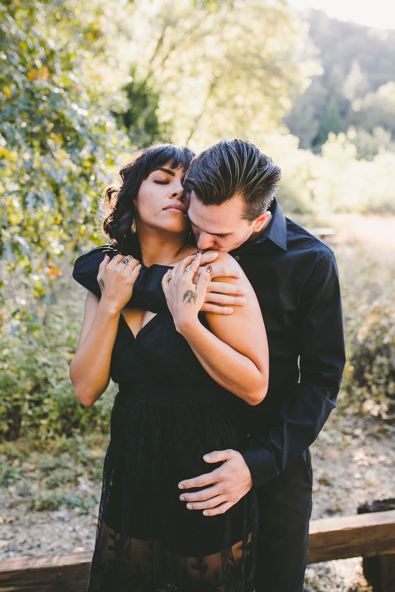 A couple in black outfits share a romantic embrace outdoors, with the man kissing the woman's shoulder.