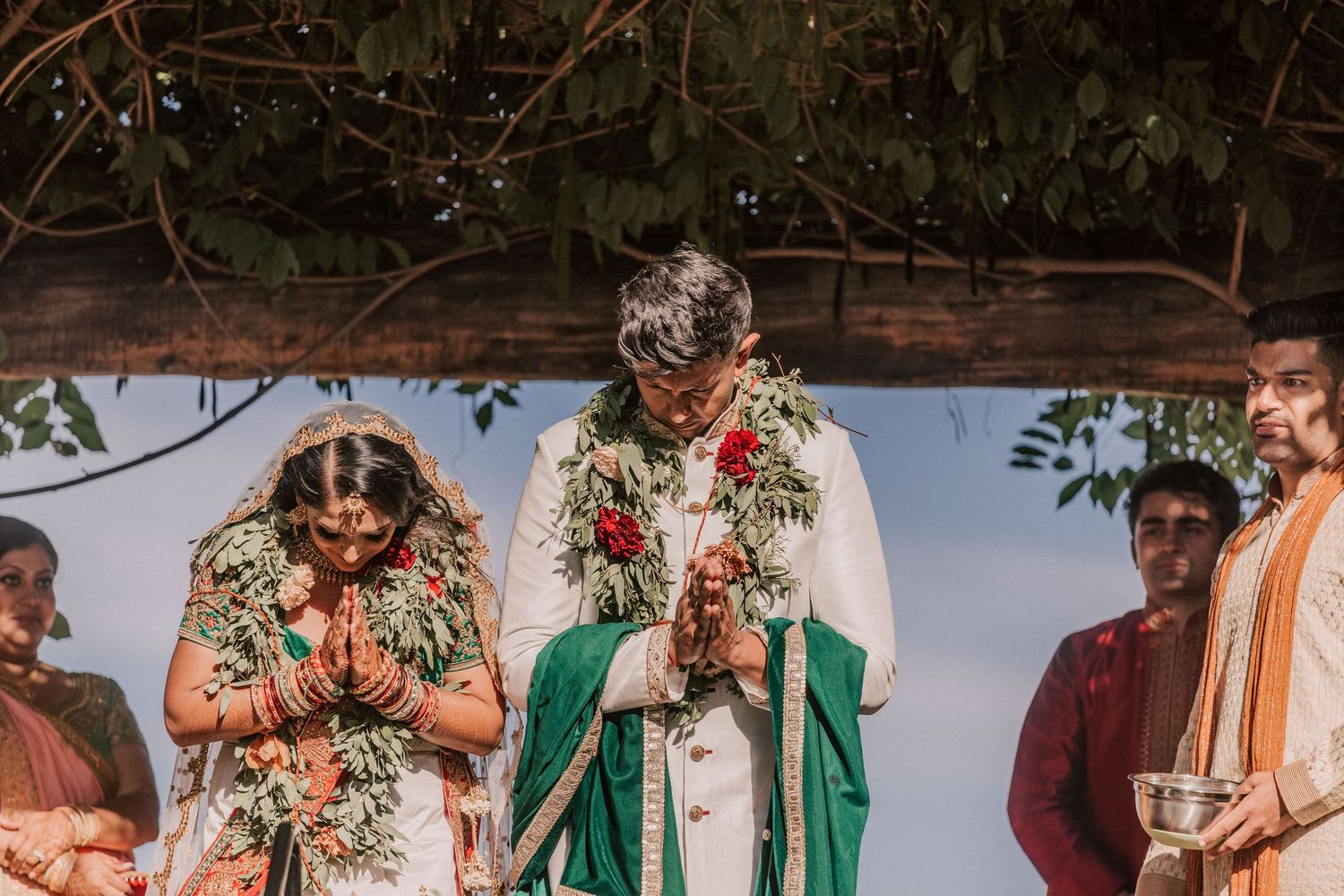Indian bride and groom in prayer pose during outdoor Hindu wedding ceremony.