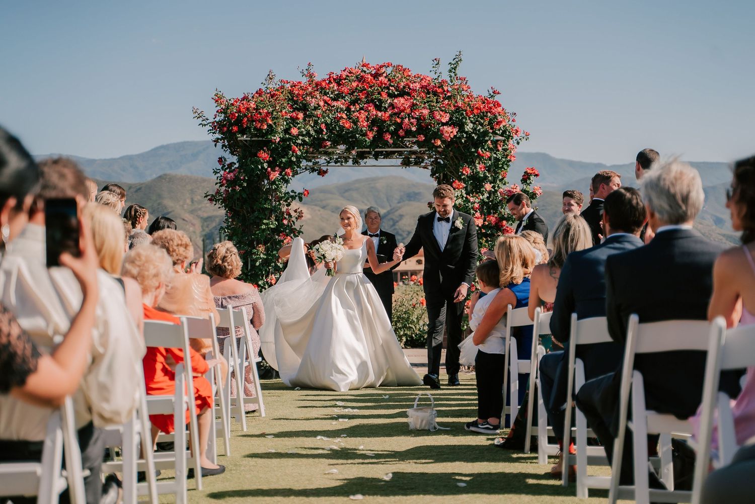 Bride and groom walk down outdoor aisle under red floral arch with mountain views and seated wedding guests.