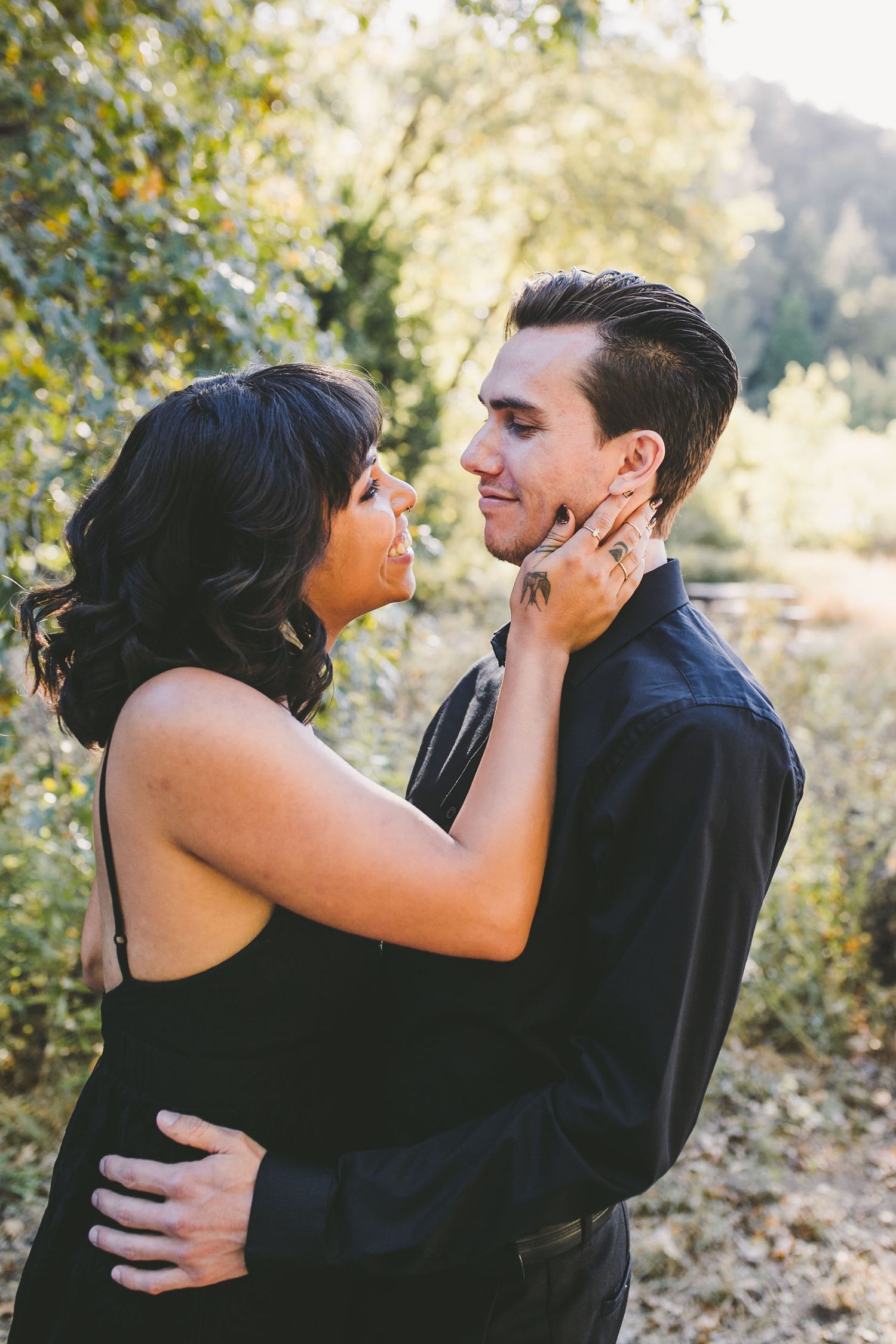 A couple in black outfits embrace outdoors, the woman tenderly touching the man's face amid lush green trees.