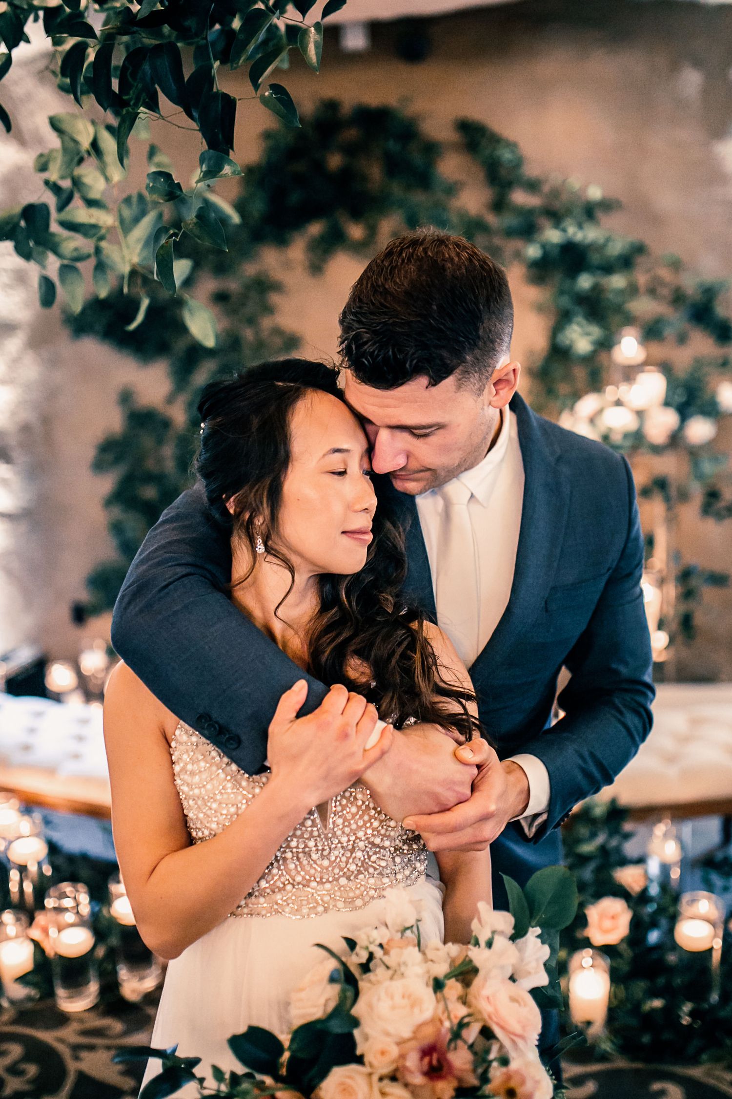 A couple shares a tender embrace at their wedding reception, surrounded by candlelight and lush greenery.