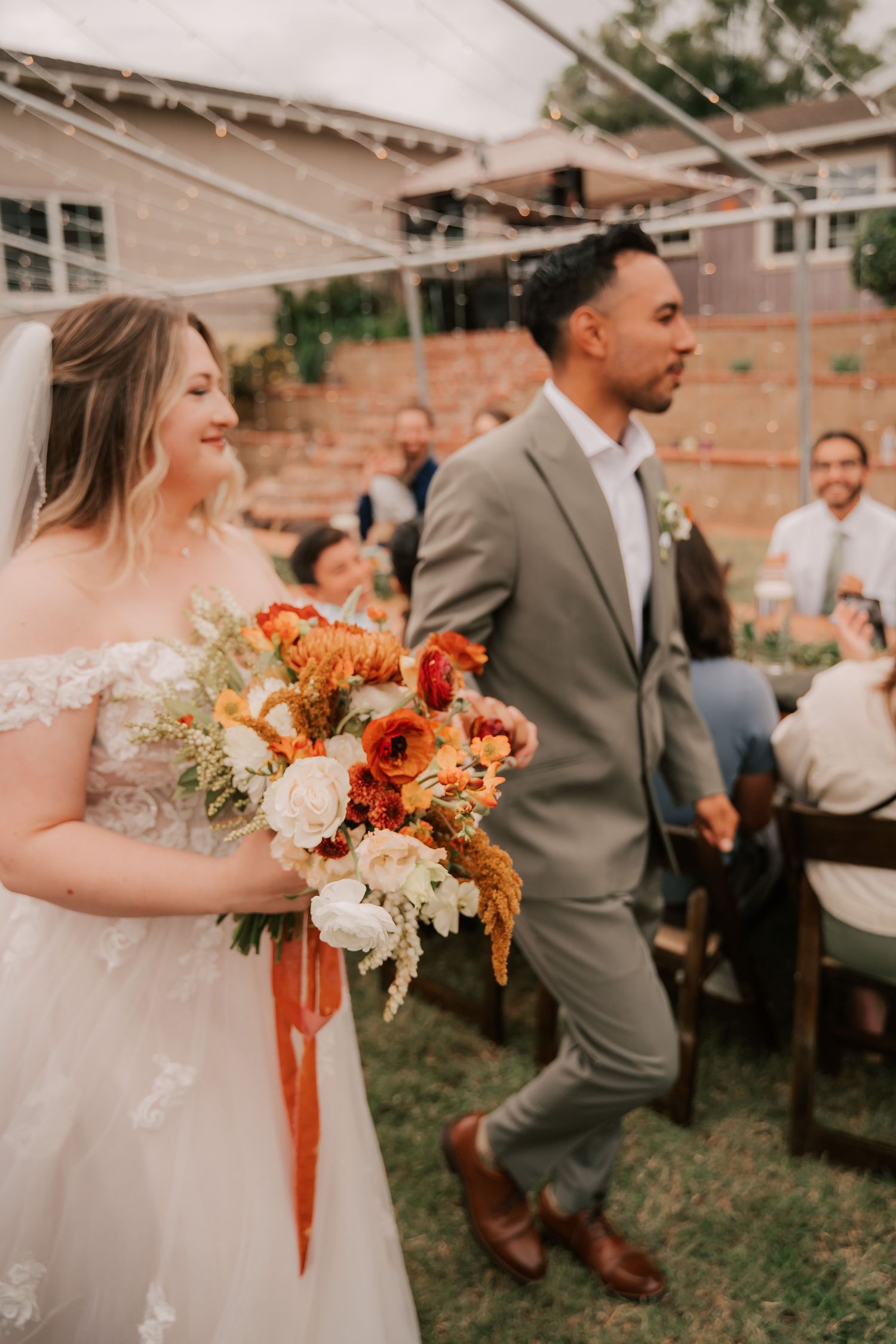 Bride holding autumn bouquet with groom in gray suit walking past guests at outdoor wedding ceremony.