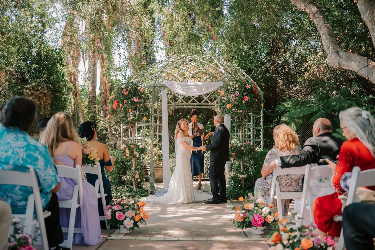 Outdoor garden wedding ceremony with bride and groom at floral arch, guests seated on white chairs amid lush greenery.