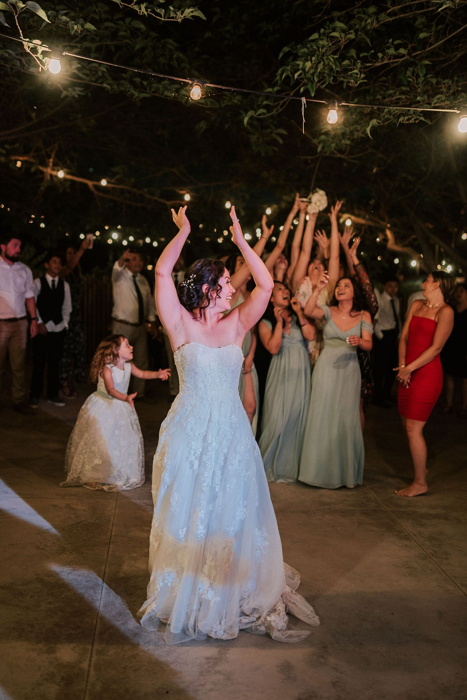 Bride tossing bouquet to excited guests at outdoor wedding reception with string lights.