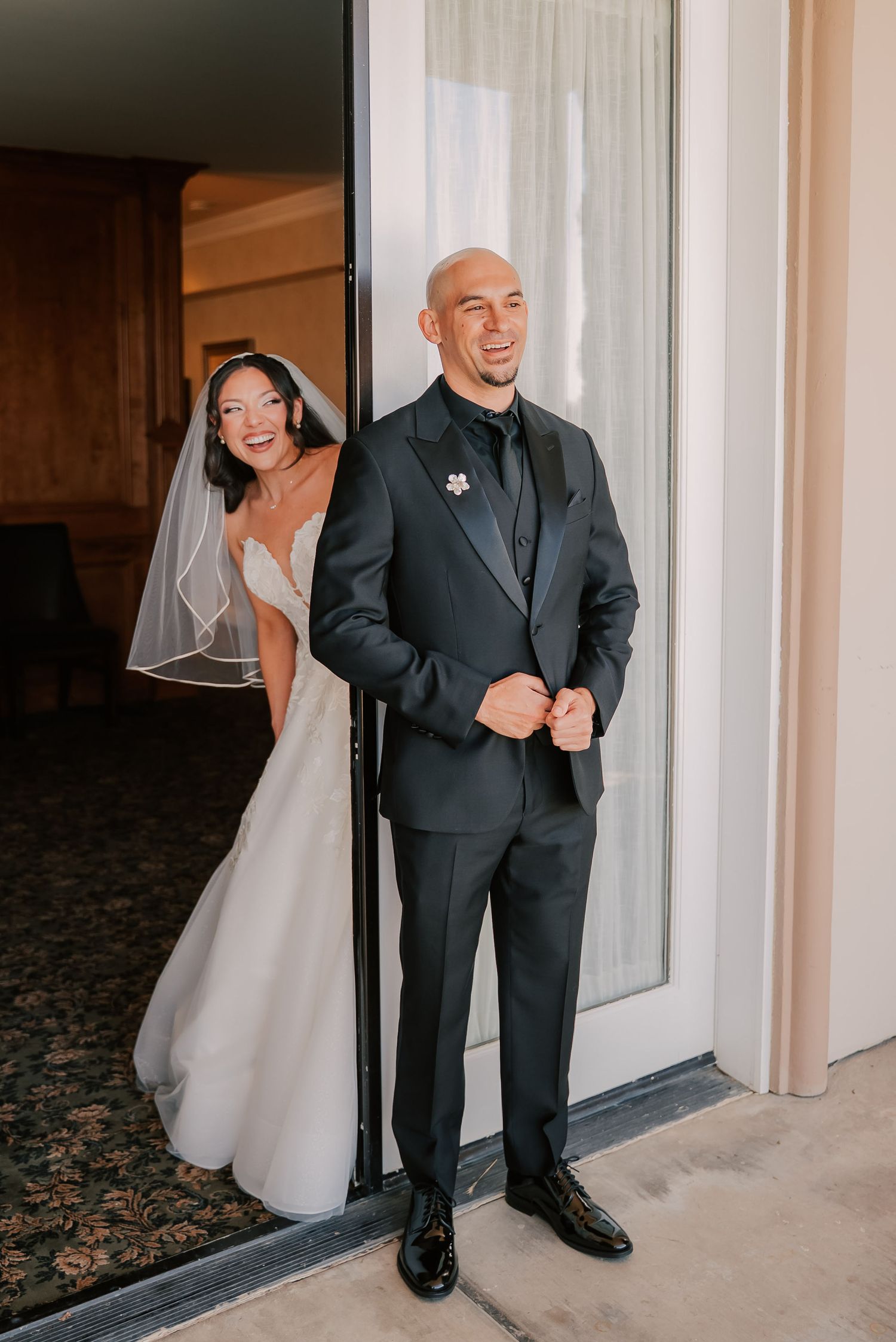 Bride in white gown and veil peeks behind groom in black tuxedo during wedding first look moment.
