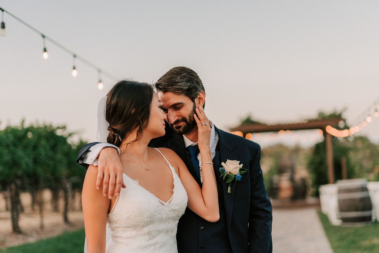 Bride and groom share a romantic moment at outdoor wedding venue with string lights at dusk.