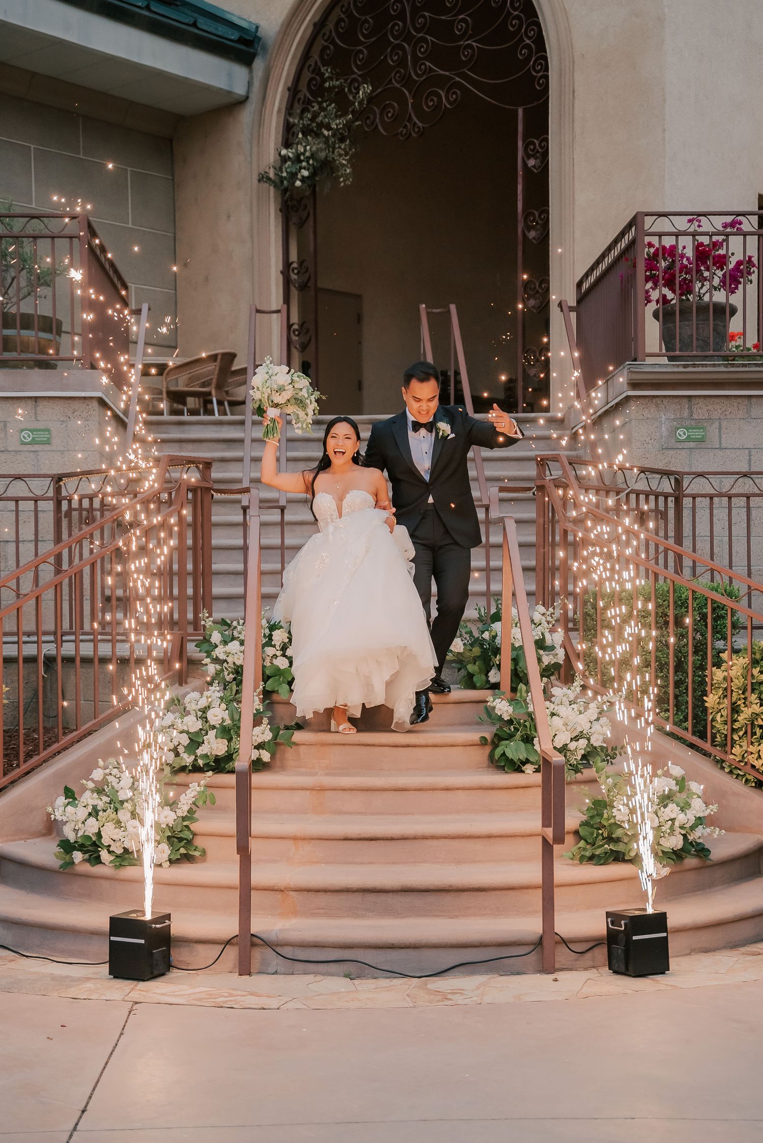 Bride and groom descend grand staircase with sparklers and white floral arrangements during elegant wedding reception exit.