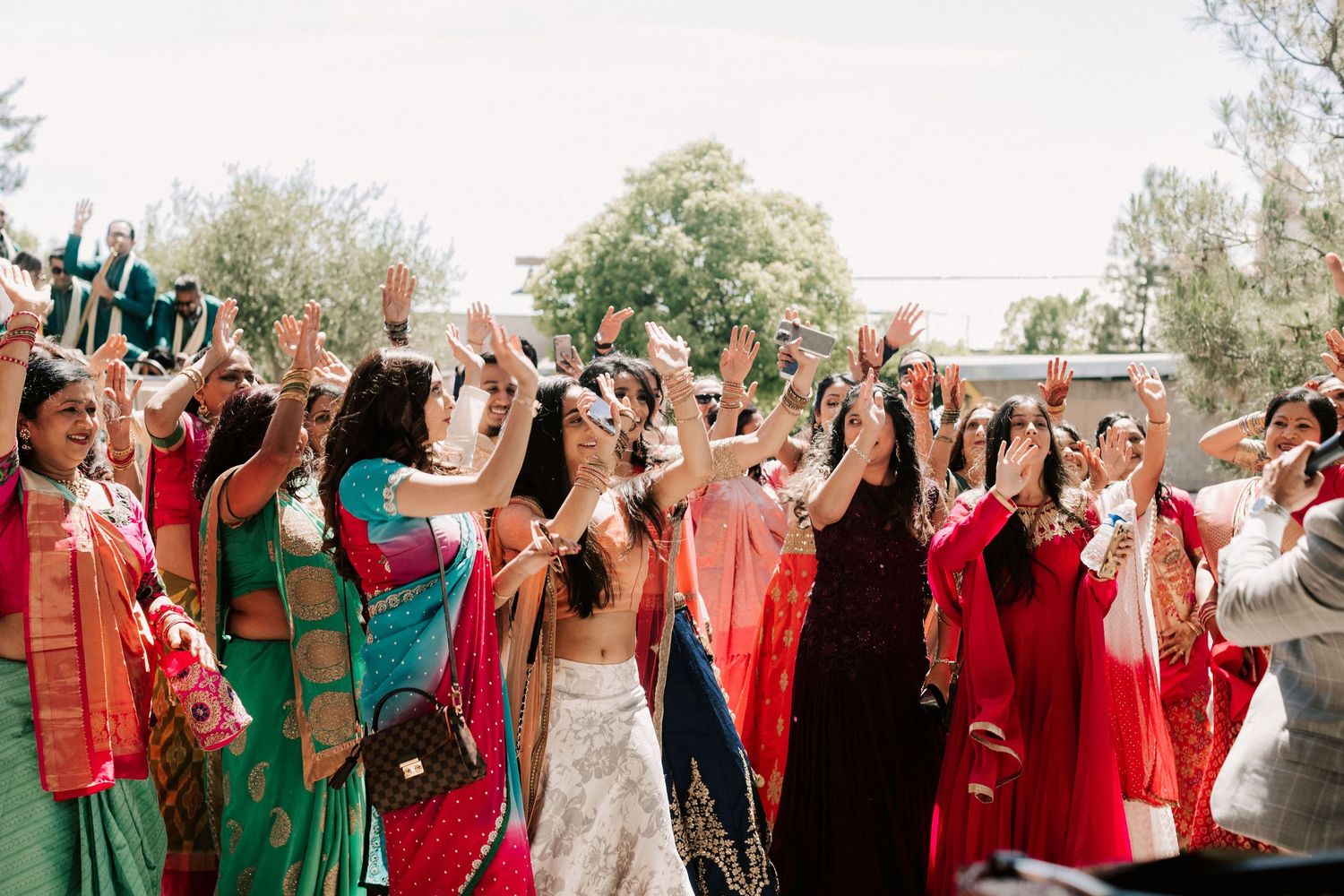 Indian wedding guests in colorful sarees celebrating outdoors with raised hands.