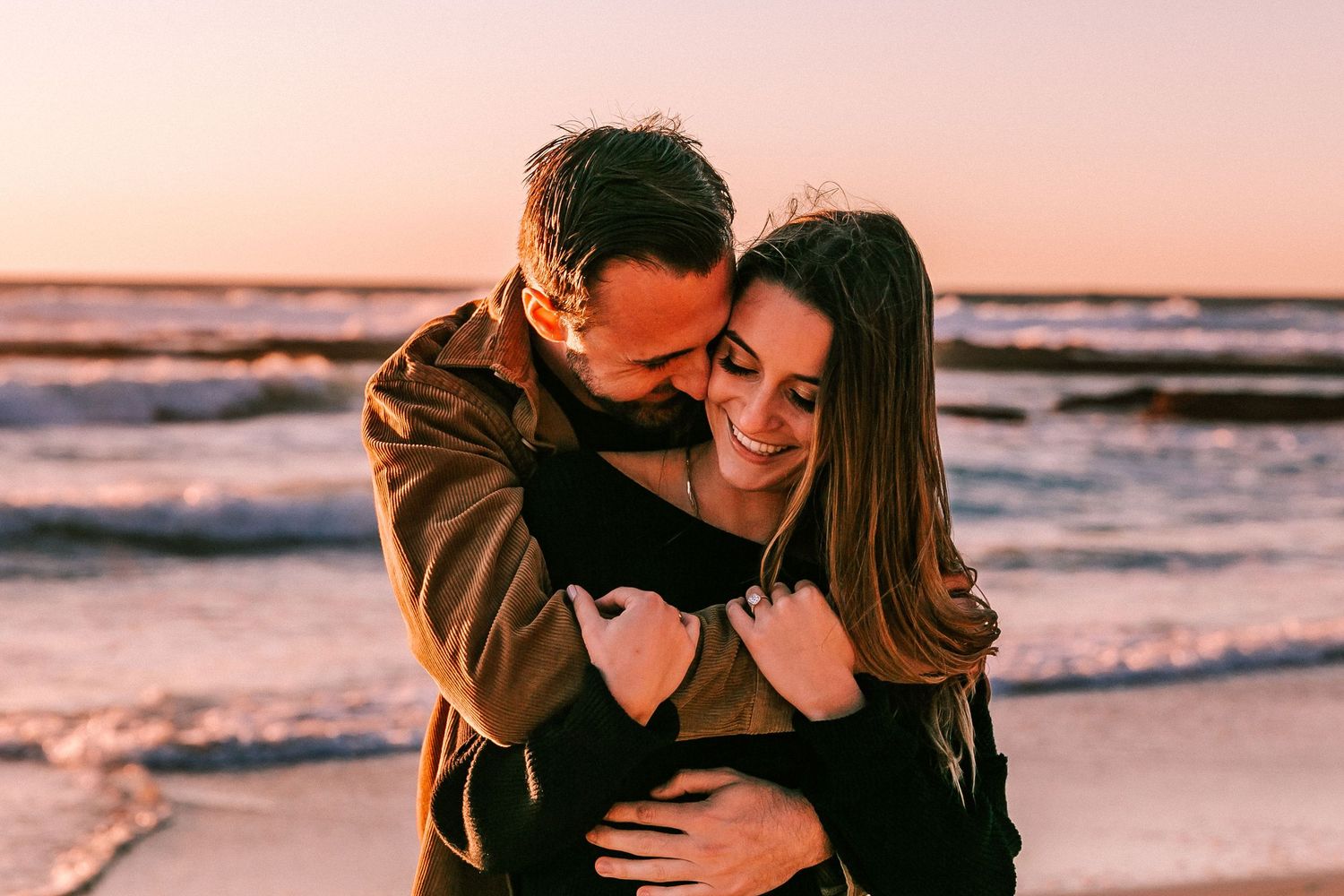 Couple sharing a warm embrace on beach at pink and orange sunset, smiling joyfully.
