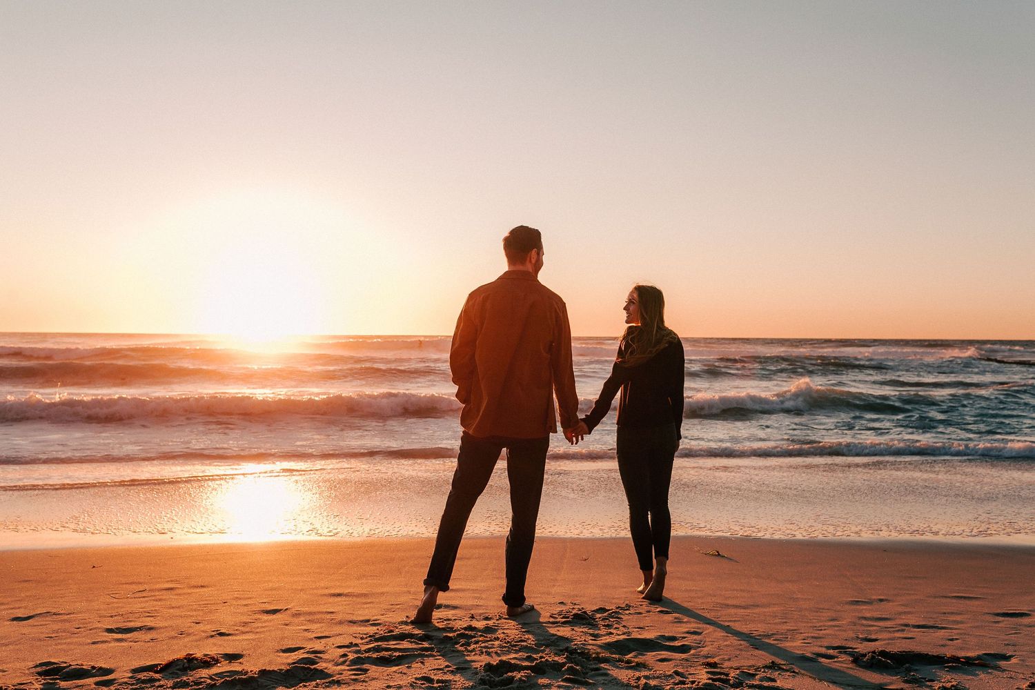 Couple holding hands on beach silhouetted against a stunning golden sunset over the ocean waves.