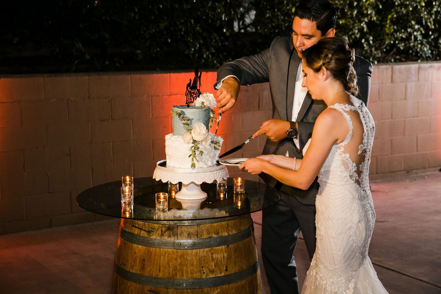 Bride and groom cutting a three-tier wedding cake displayed on a rustic wine barrel at an outdoor evening reception.