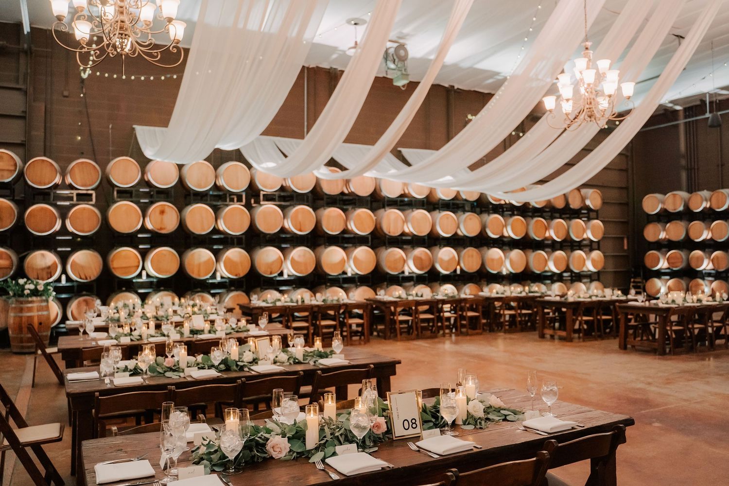 Elegant winery wedding reception with barrel wall backdrop, draped ceilings, chandeliers, and candlelit wooden tables with greenery.