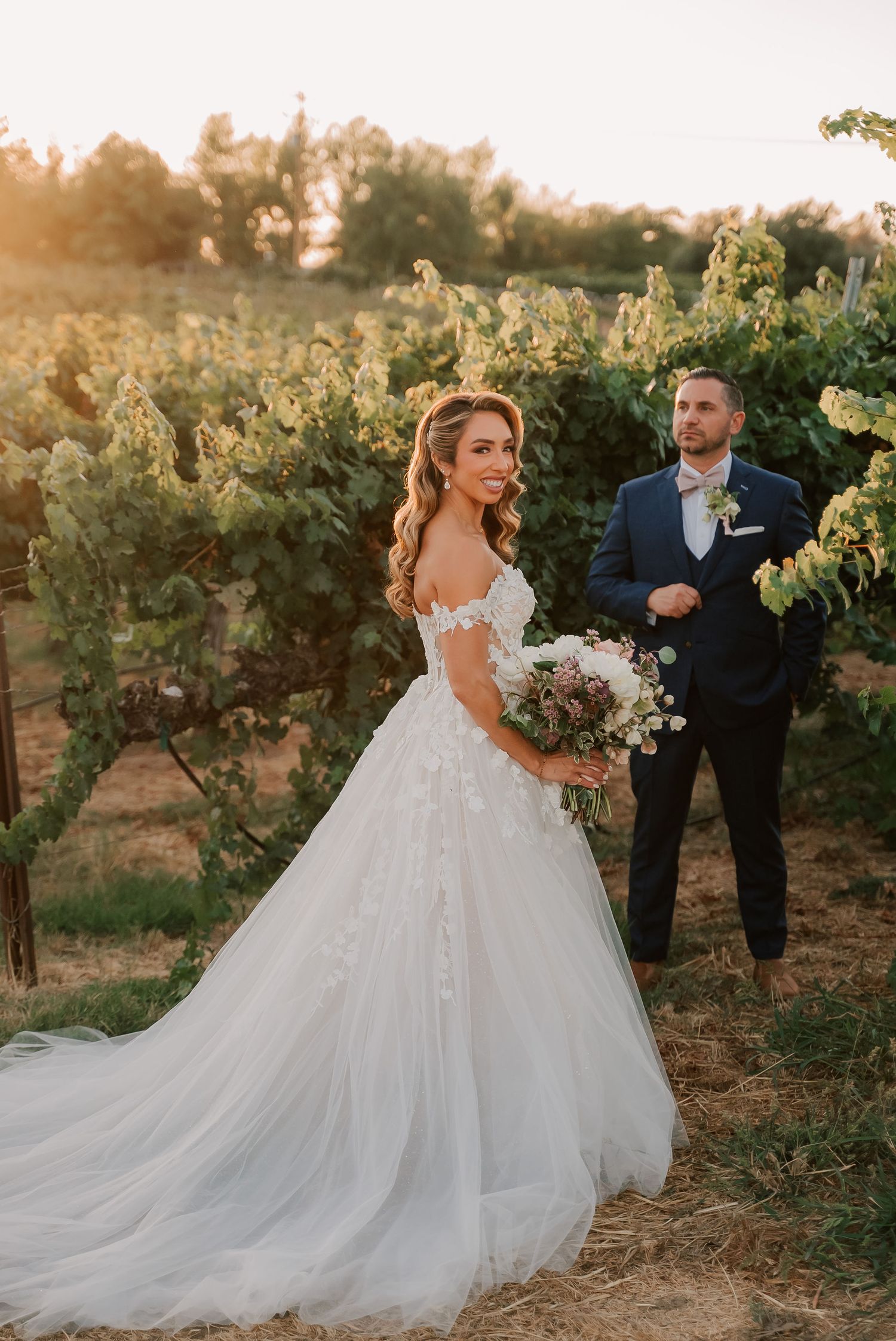 Bride in lace ball gown holds bouquet in vineyard at golden hour, groom in navy suit stands behind her.