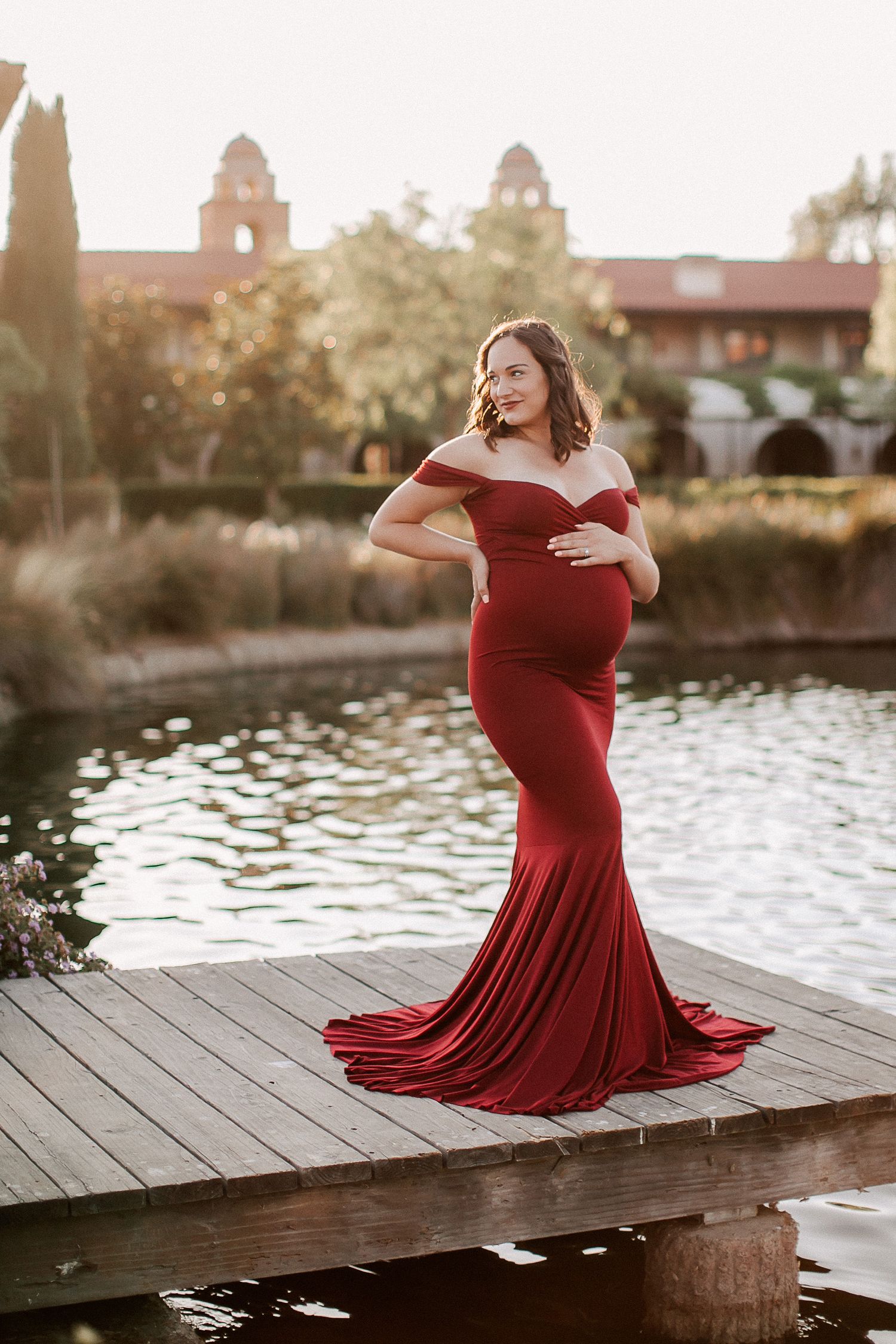 Pregnant woman in elegant red off-shoulder mermaid gown poses on wooden dock by serene pond at golden hour.