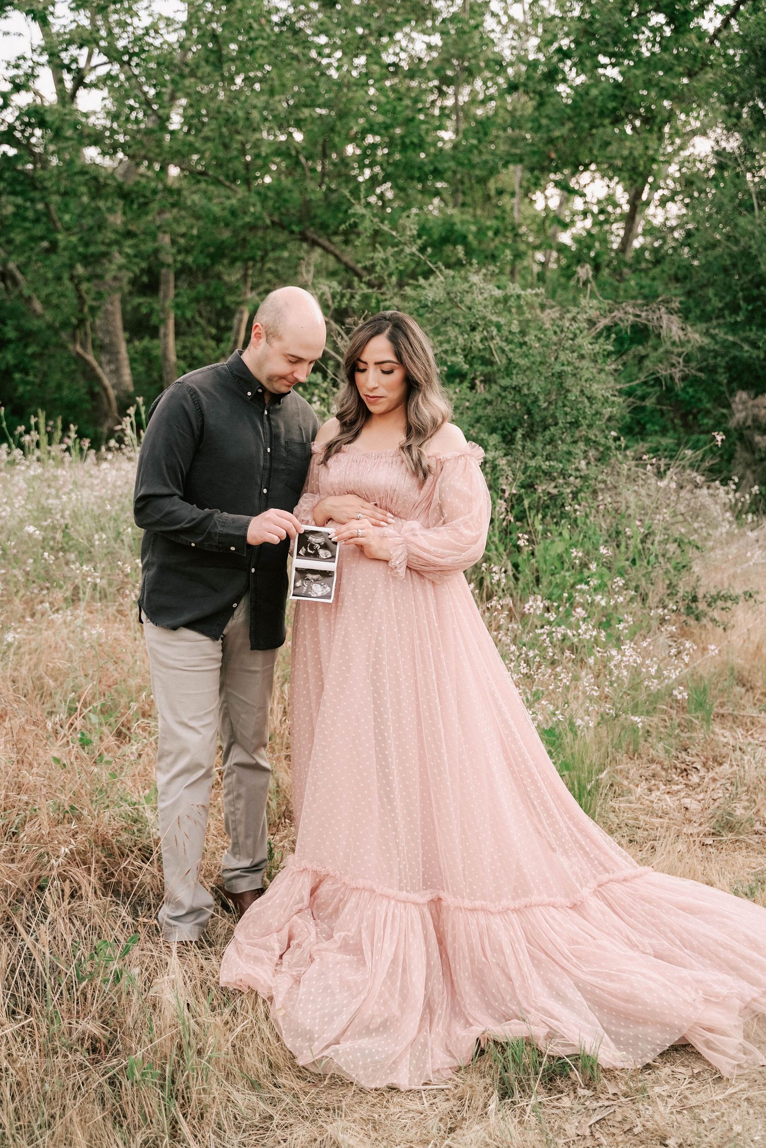 Couple in a field holding an ultrasound photo, woman in pink maternity gown, man in black shirt, green trees background.