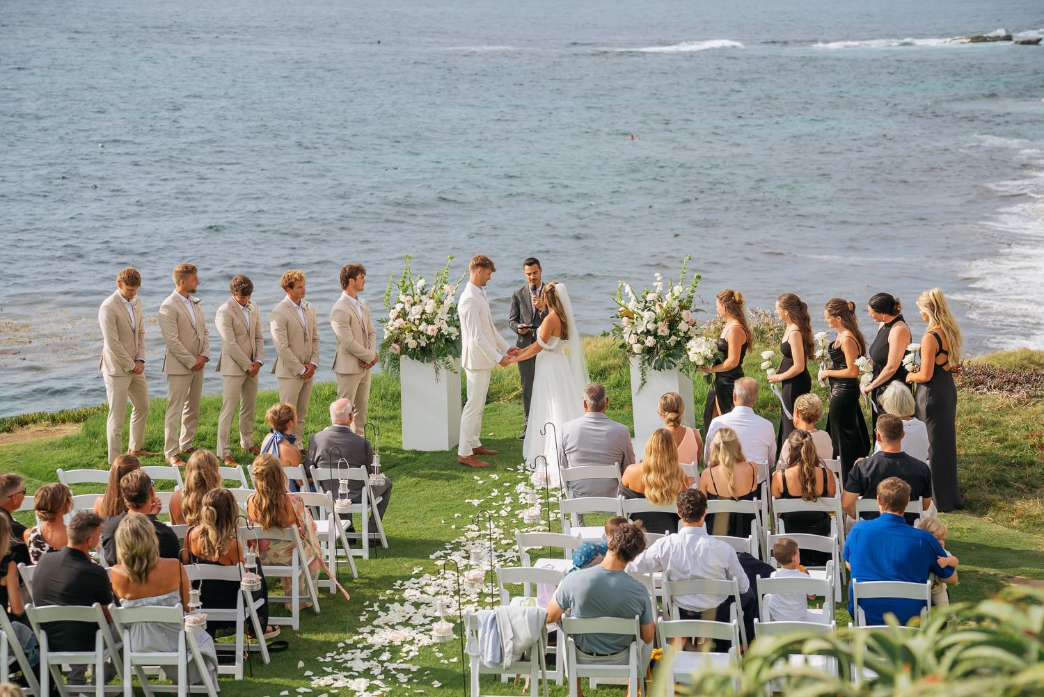 Elegant oceanside wedding ceremony on green lawn with white chairs, floral arrangements, wedding party, and sea views.