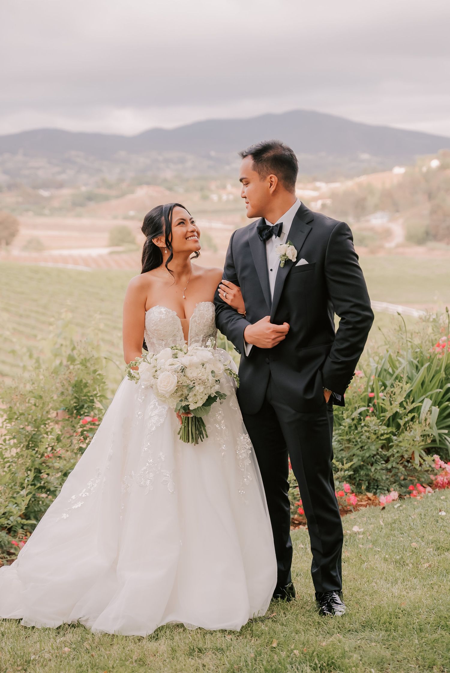 Bride in strapless lace gown holding white bouquet stands with groom in black tuxedo at vineyard wedding venue.