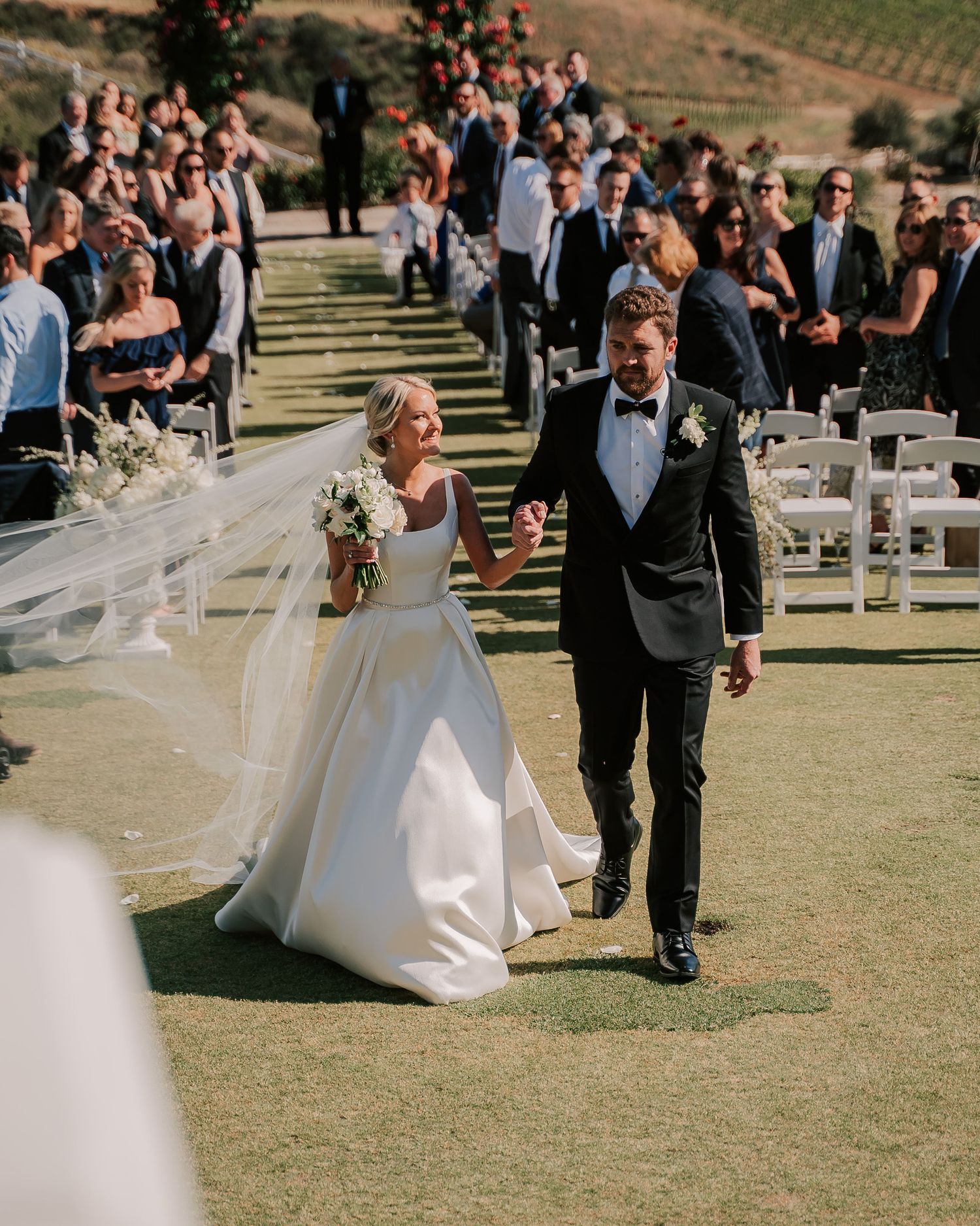 Bride and groom walk down the aisle at an outdoor wedding ceremony on a sunny day.