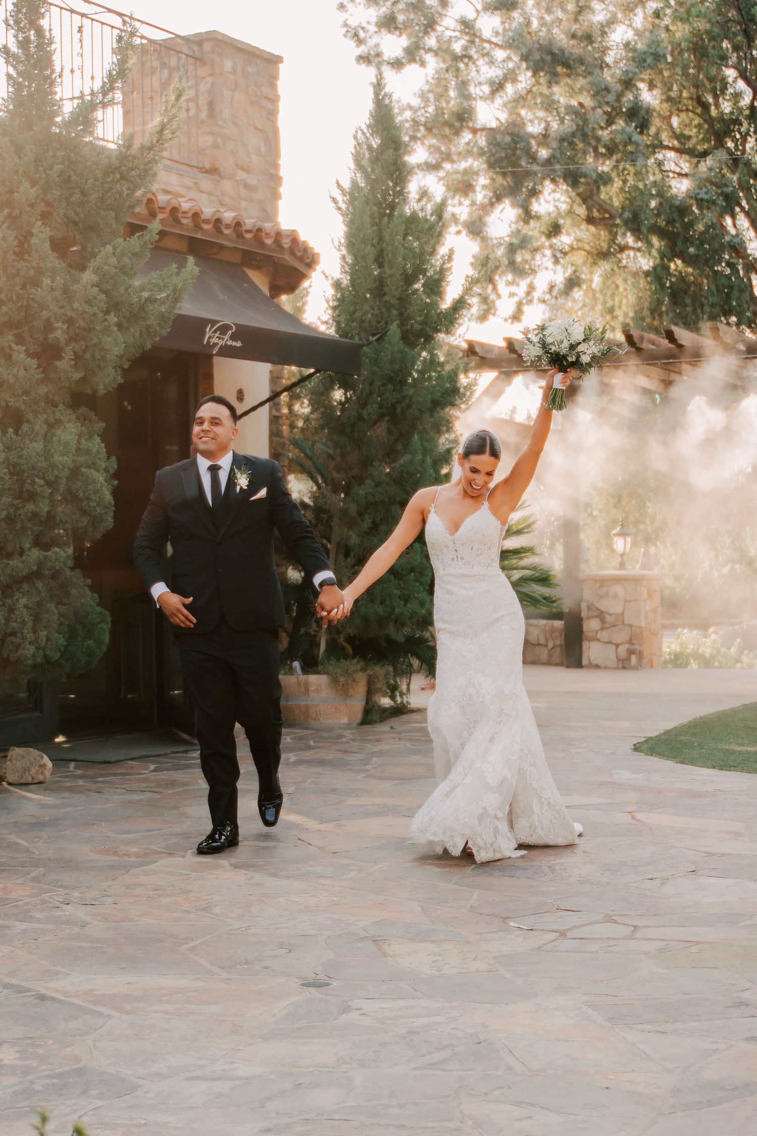 Bride raising bouquet and groom in tuxedo celebrate after wedding ceremony at elegant outdoor venue.
