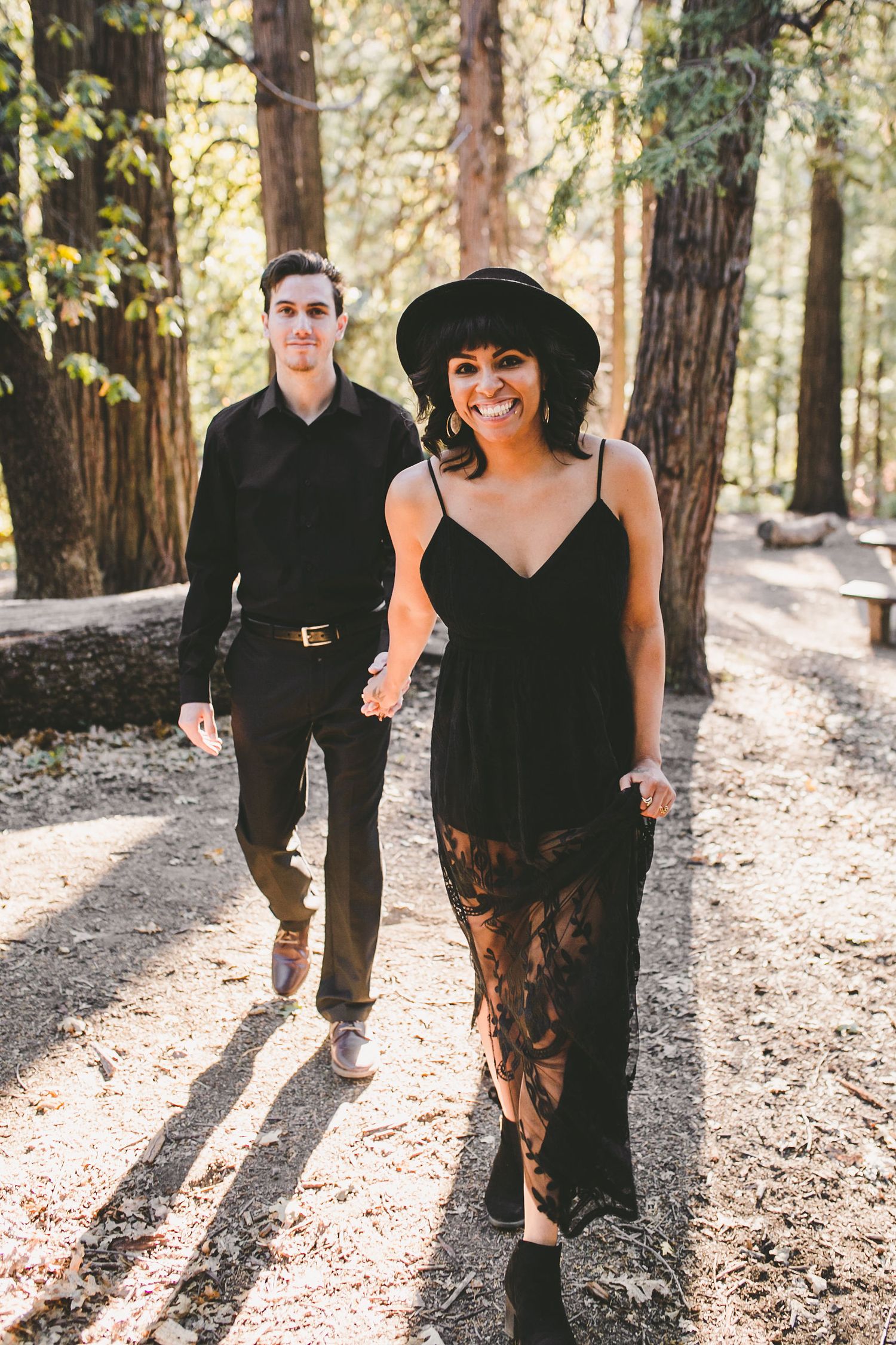 Couple in black outfits walking hand-in-hand through a sunlit forest, woman smiling in a wide-brim hat.