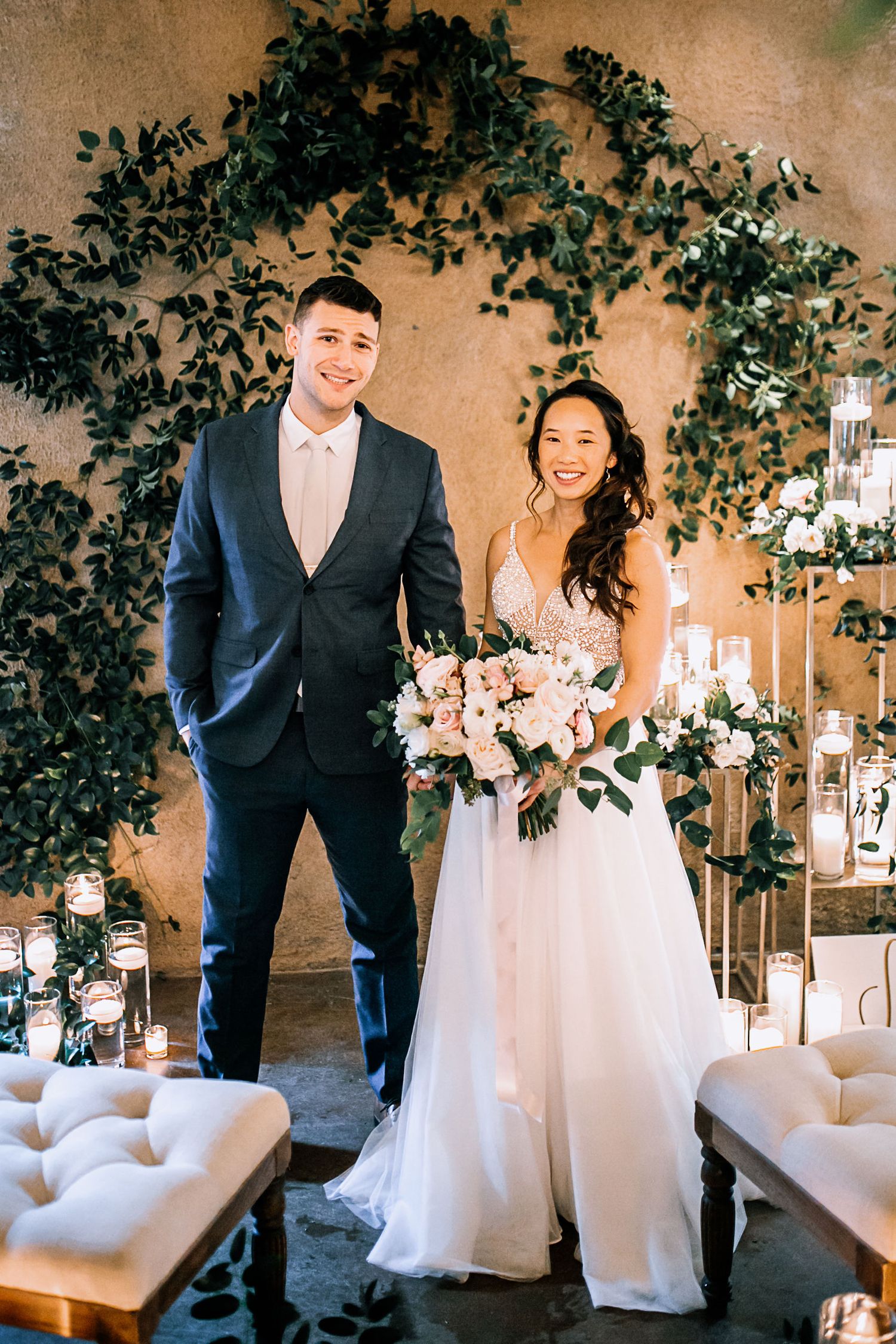 Bride and groom smiling at wedding altar with lush greenery arch, white floral bouquet, and candlelit decor.