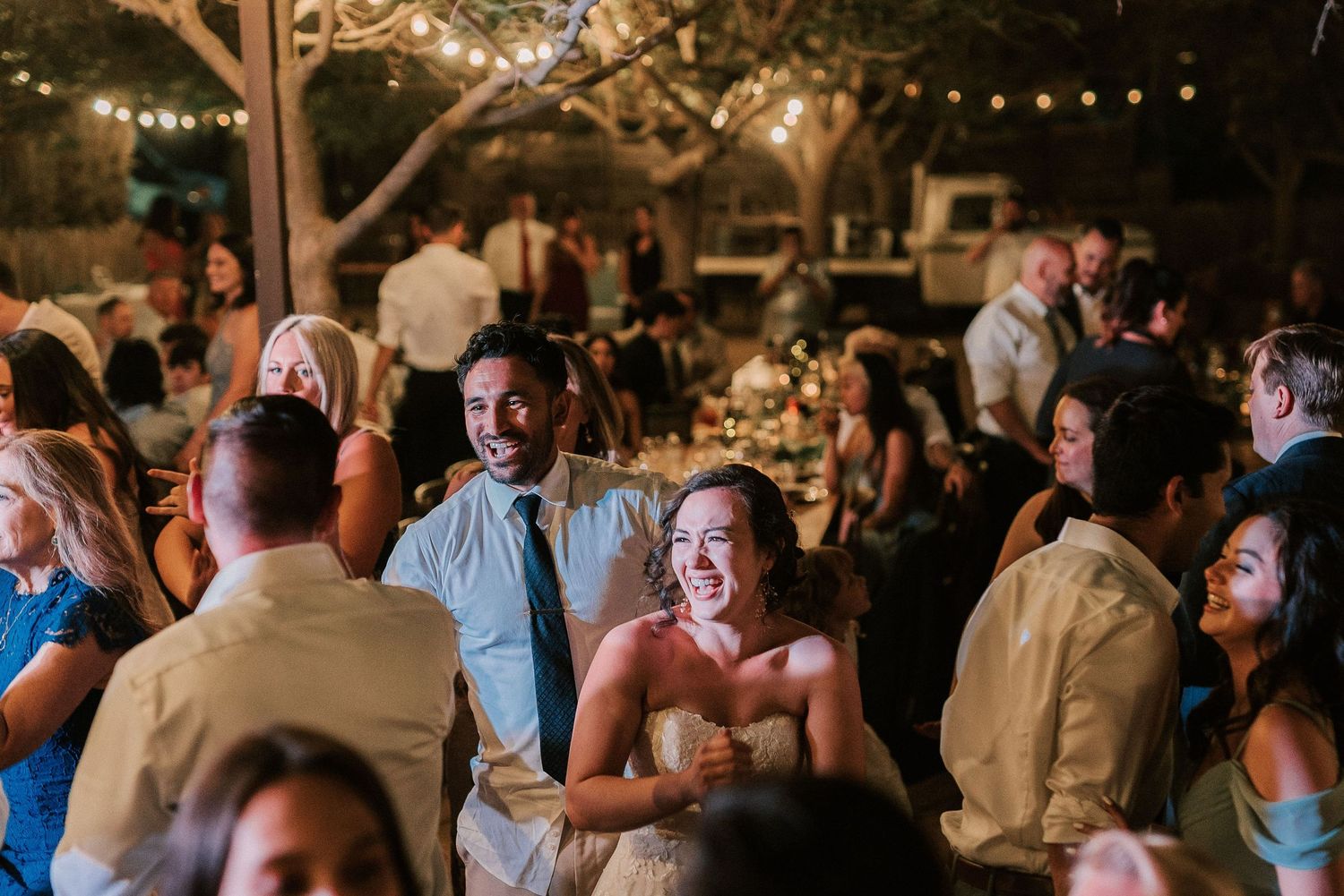 Bride and groom laughing joyfully with guests at an outdoor evening wedding reception under string lights.
