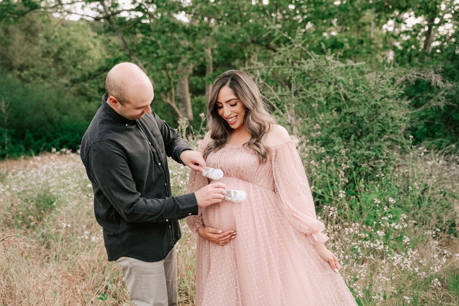 Couple in outdoor maternity photoshoot, woman in pink gown holding baby shoes over baby bump.
