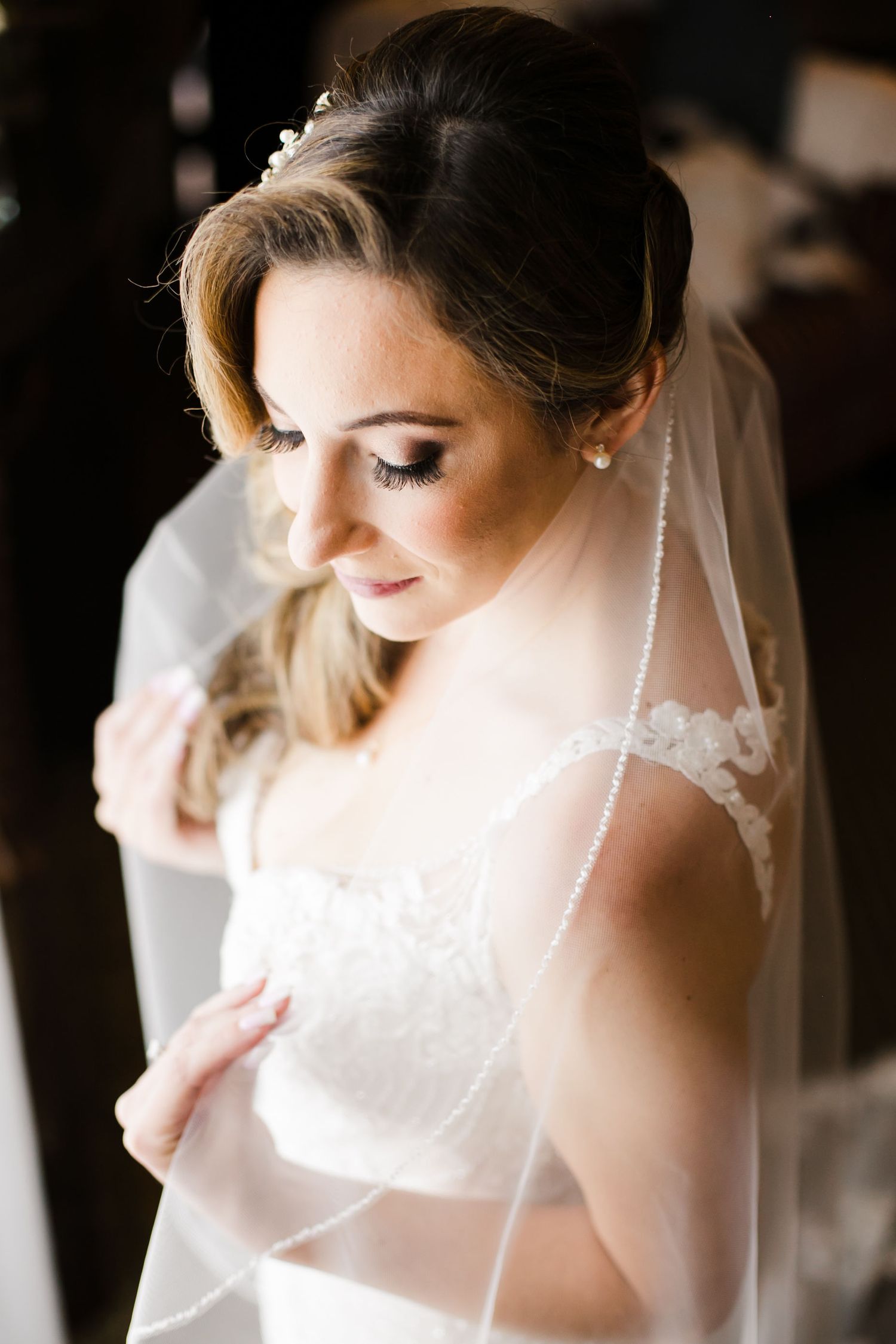 Elegant bride in lace wedding dress and pearl-trimmed veil, wearing pearl earrings and hair accessory, looking down gracefully.