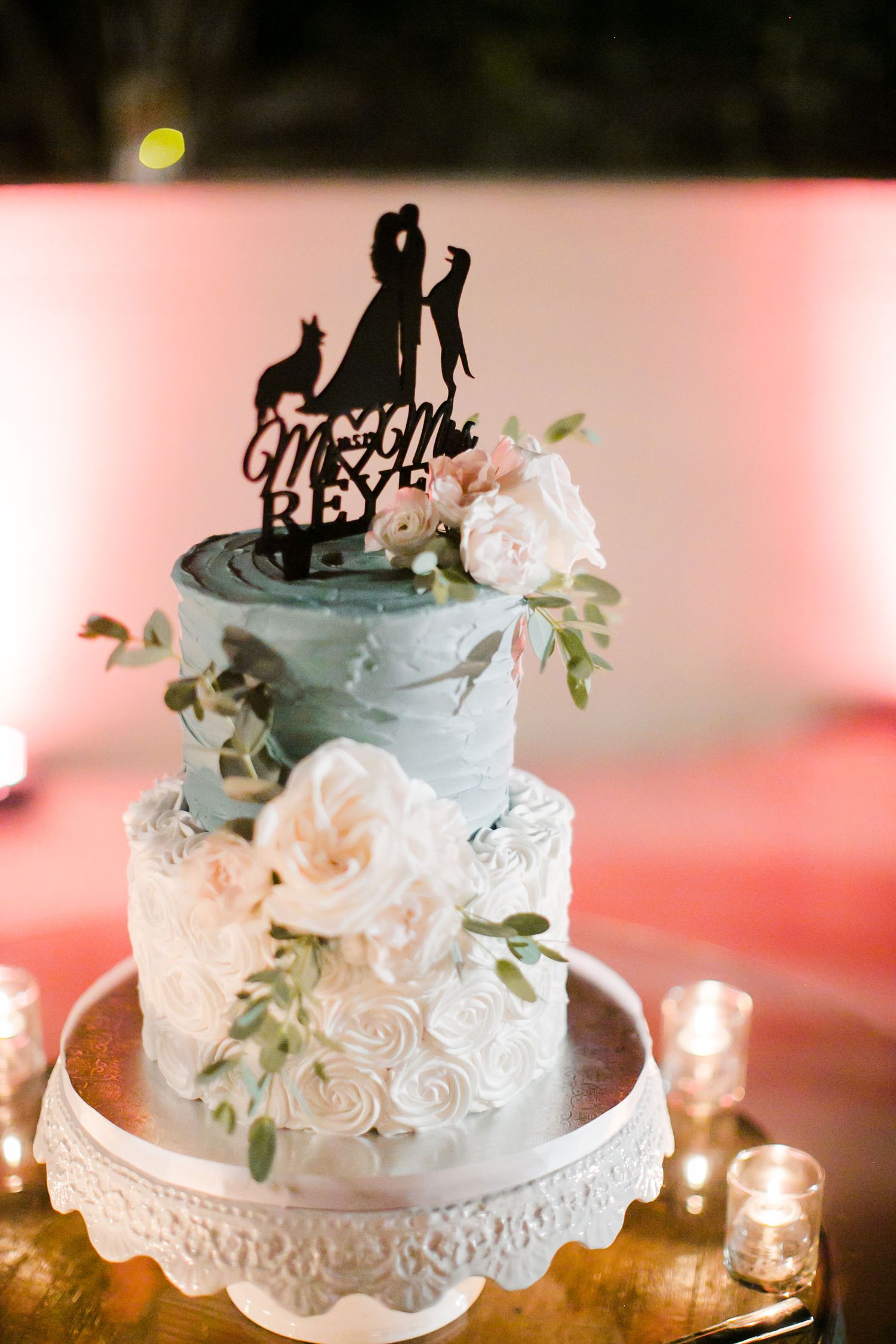 Two-tier blue and white wedding cake with floral decorations and silhouette topper reading Mr. and Mrs. Rey, surrounded by candles.
