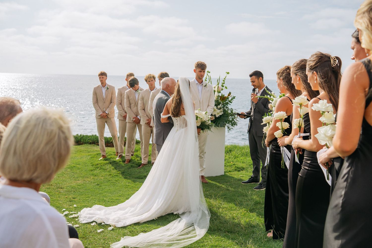 Outdoor wedding ceremony by the ocean with bride, groom, groomsmen in tan suits, and bridesmaids in black dresses.