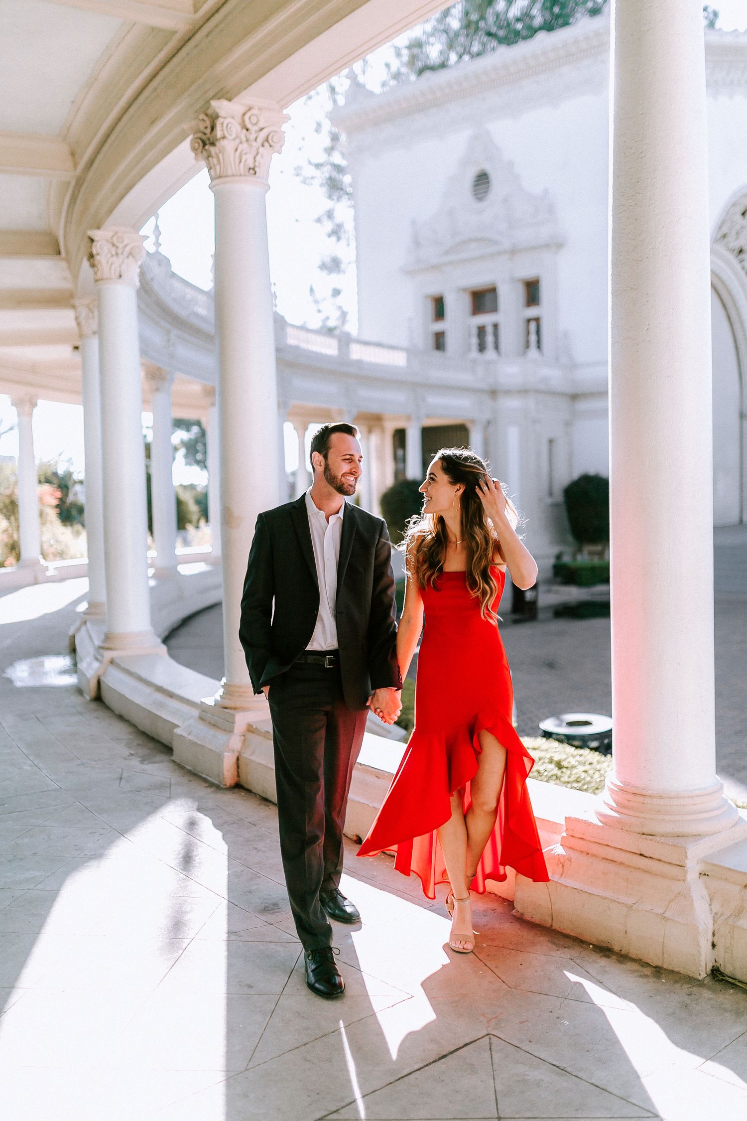 Couple holding hands walking through white columned archway, woman wearing flowing red dress.