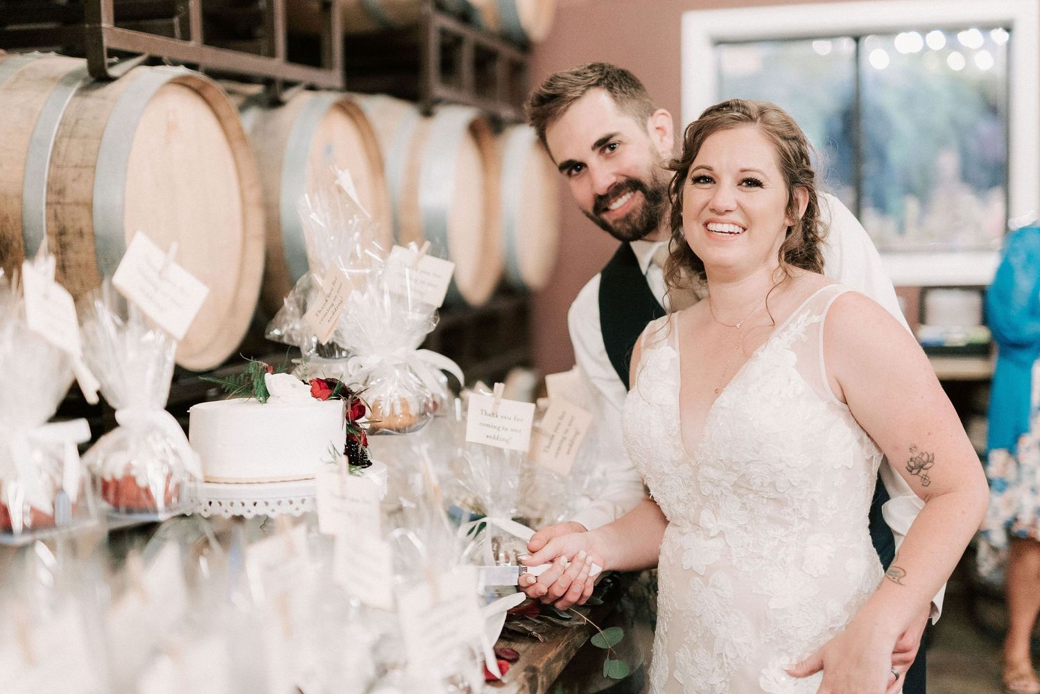 Bride and groom smiling while cutting wedding cake at a winery reception with wooden barrels in background.