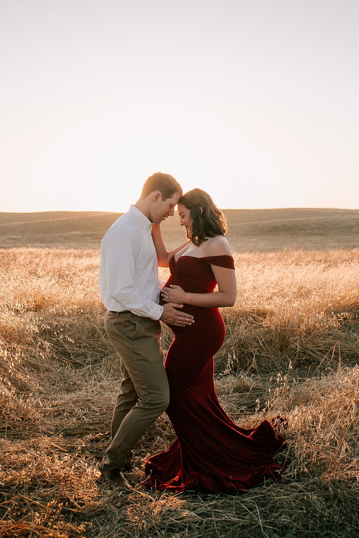 Couple sharing a tender maternity moment at golden hour in a field, woman in elegant burgundy gown.