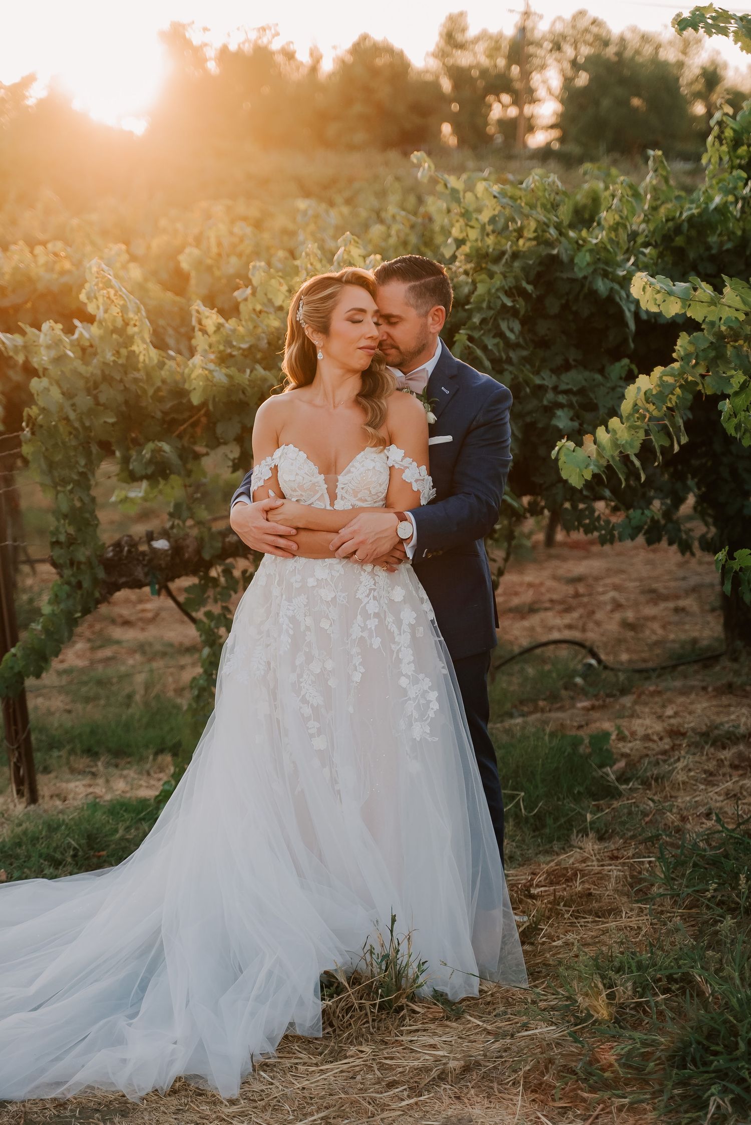 Bride and groom embrace at golden hour in a vineyard, bride wearing a floral lace off-shoulder gown with a flowing train.