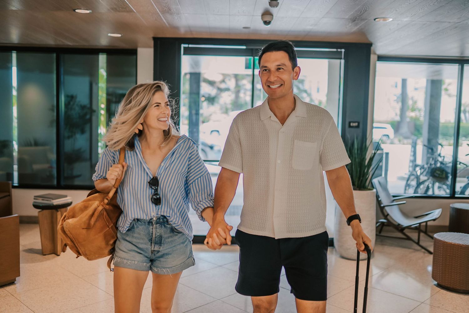 A smiling couple holds hands while walking through a bright hotel lobby, the woman carrying a brown bag and the man rolling luggage.
