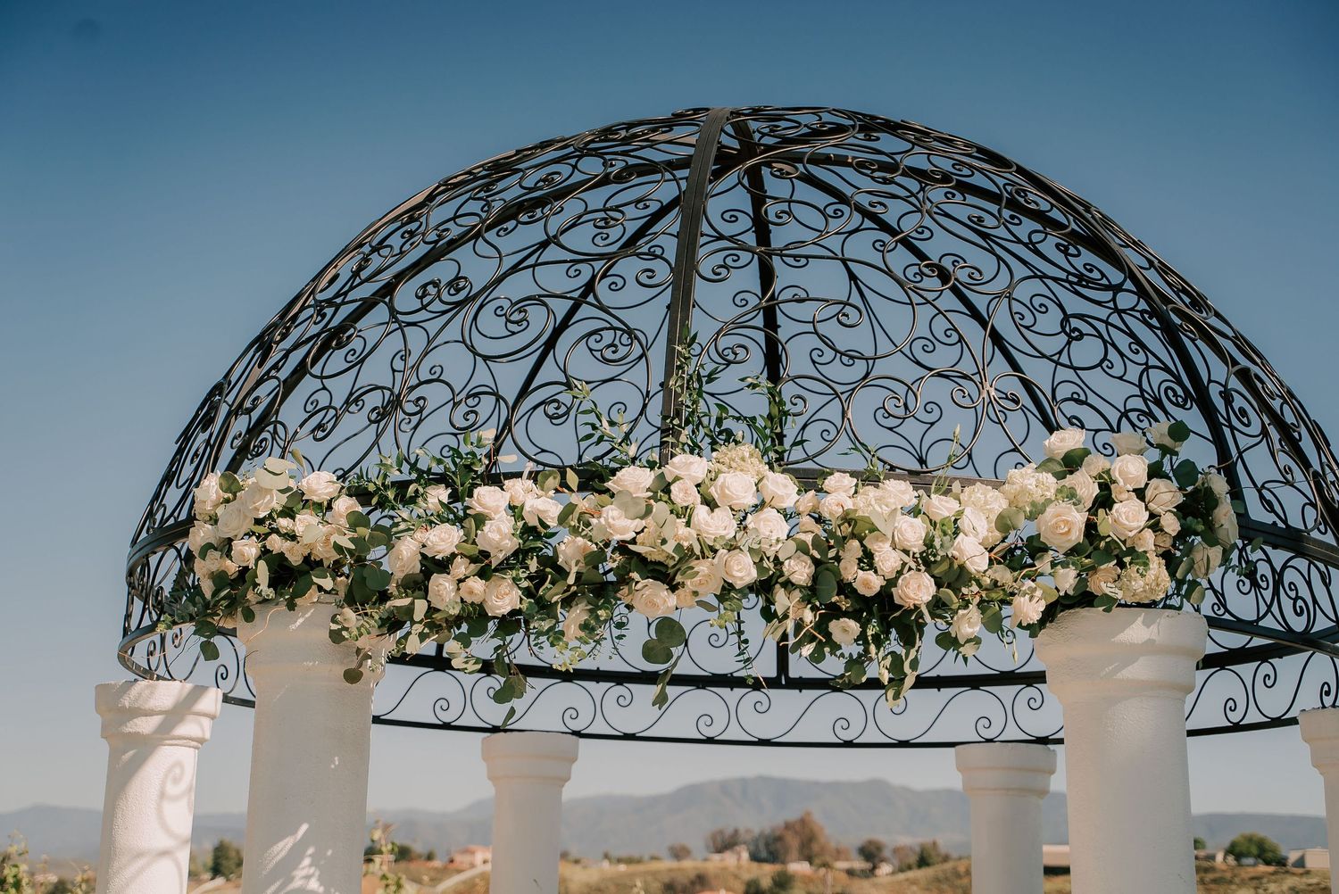 Elegant outdoor wedding gazebo with ornate black iron dome adorned with lush white roses and greenery floral arrangements.