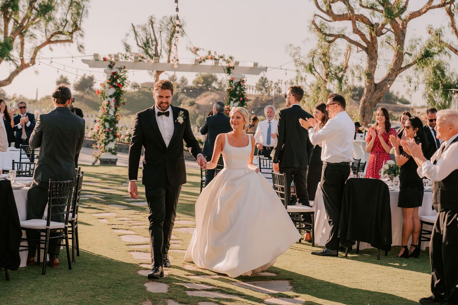 Bride and groom walk hand-in-hand down the aisle as guests applaud at an outdoor wedding reception.