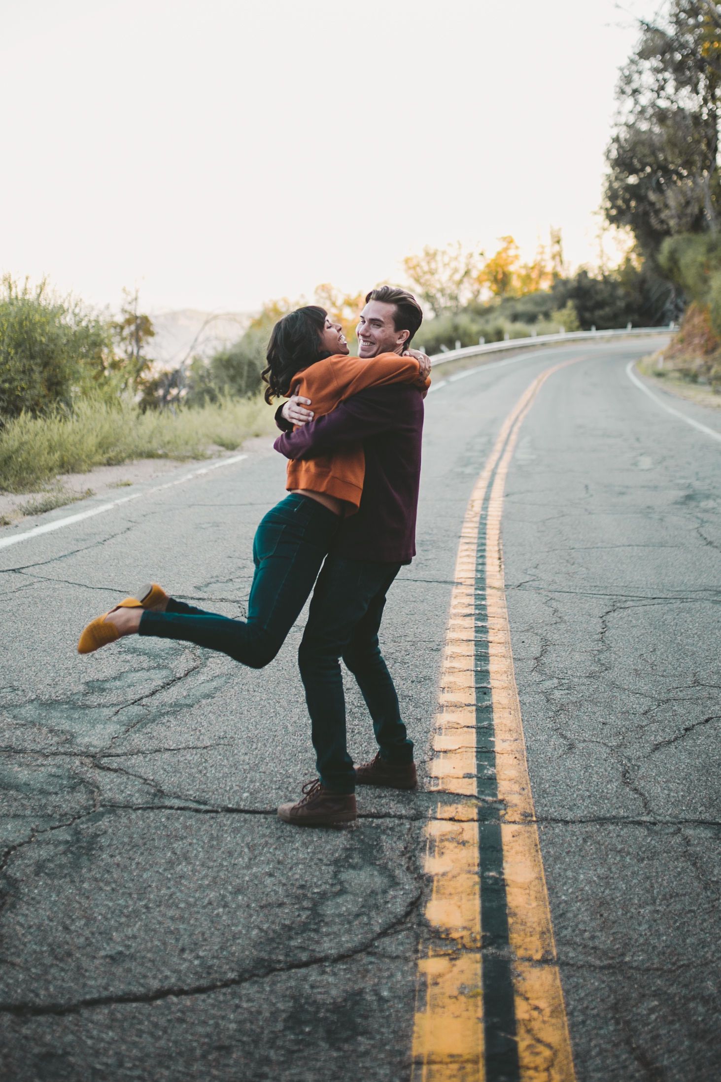Couple embracing and spinning joyfully on a winding road surrounded by lush greenery at sunset.