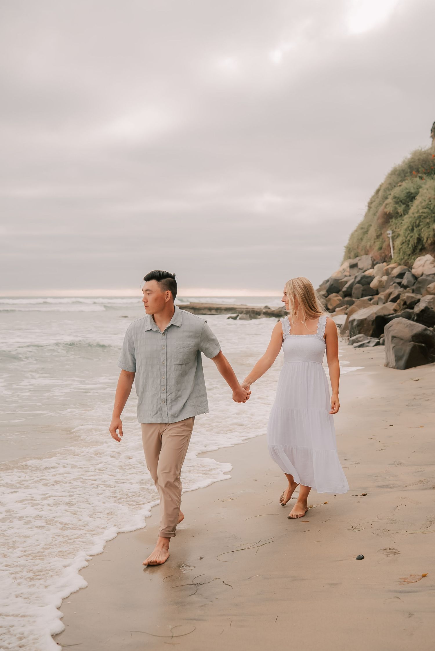 Couple holding hands walking along a sandy beach with rocky cliffs and ocean waves in the background.