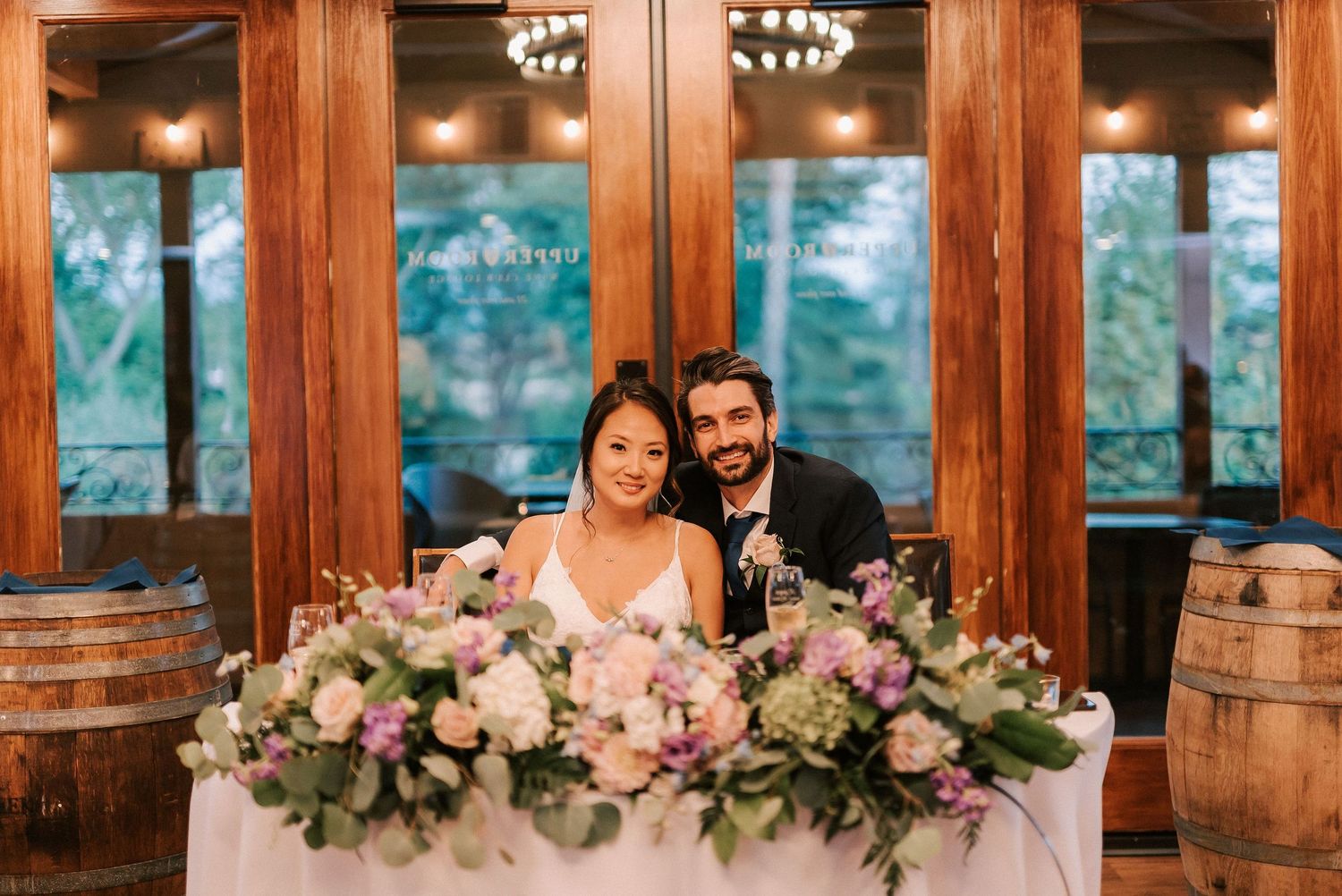 Bride and groom smiling at sweetheart table with pink floral arrangements in rustic wood venue.