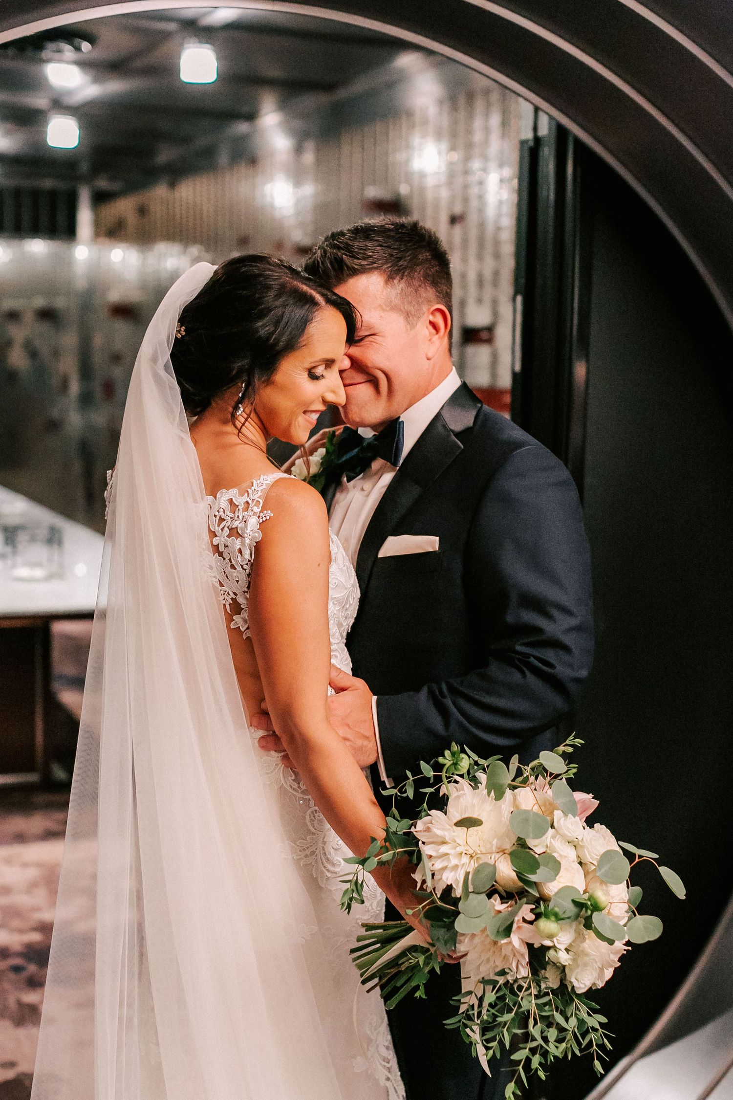 Bride and groom share an intimate moment framed by a circular mirror, bride holding a white floral bouquet.