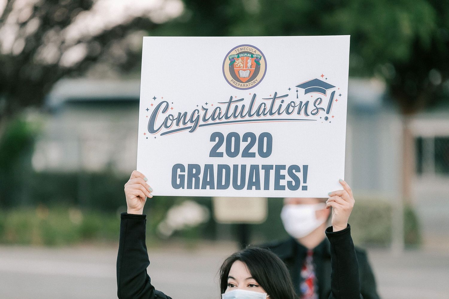 Person holding a Congratulations 2020 Graduates sign with school logo at outdoor graduation event.