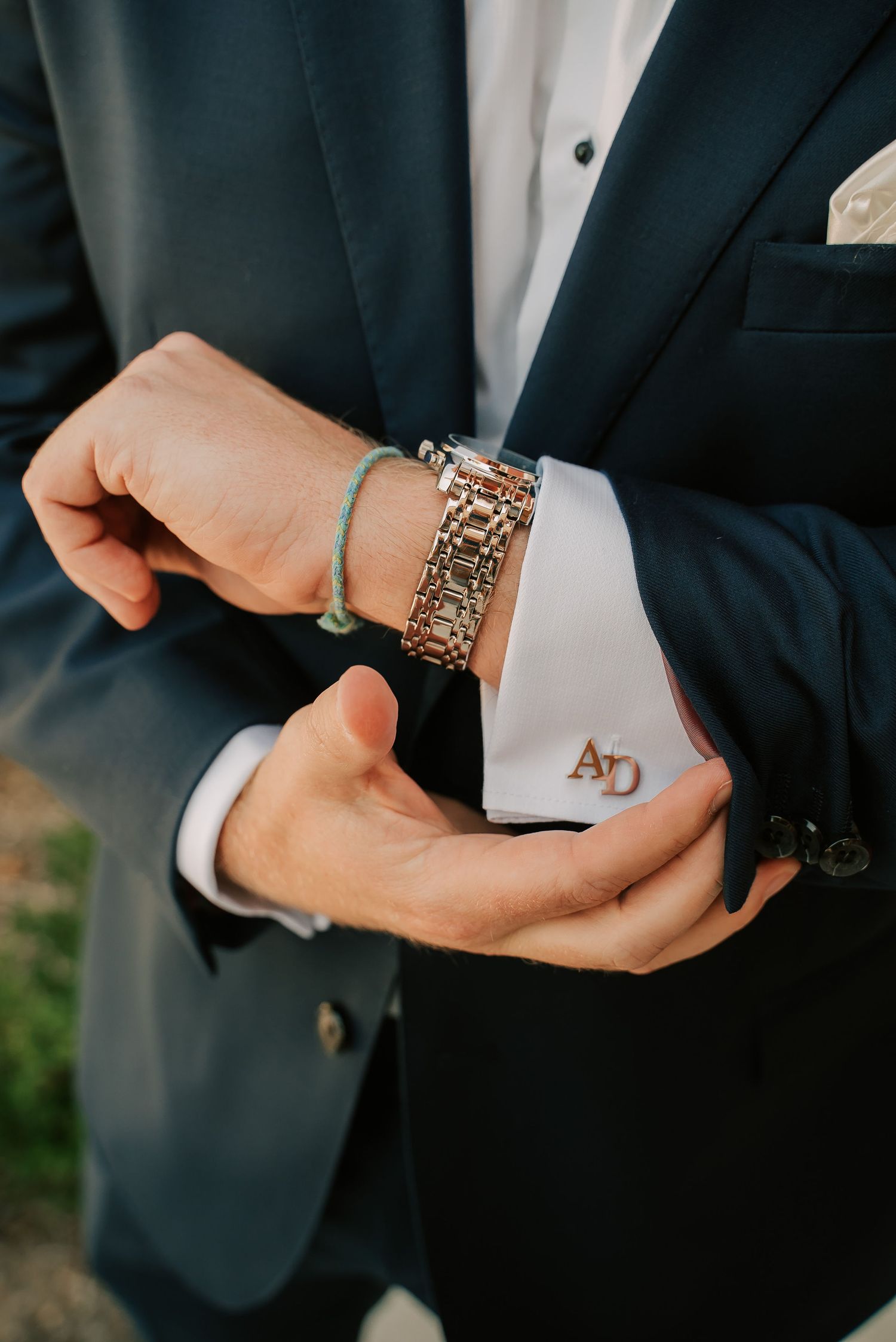 Groom adjusting monogrammed cufflinks on white dress shirt, wearing navy suit with gold bracelet and green string bracelet.